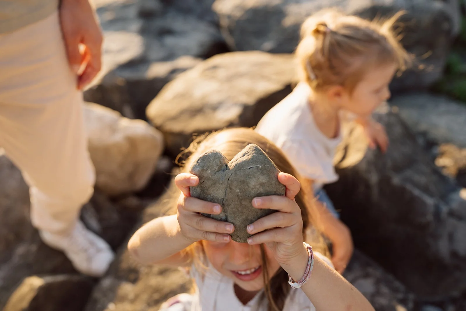 Instant spontané au bord du lac en Suisse : une enfant montre une pierre en forme de cœur lors d’une séance photo famille lifestyle, capturée avec douceur et naturel par Nonante Émotions.