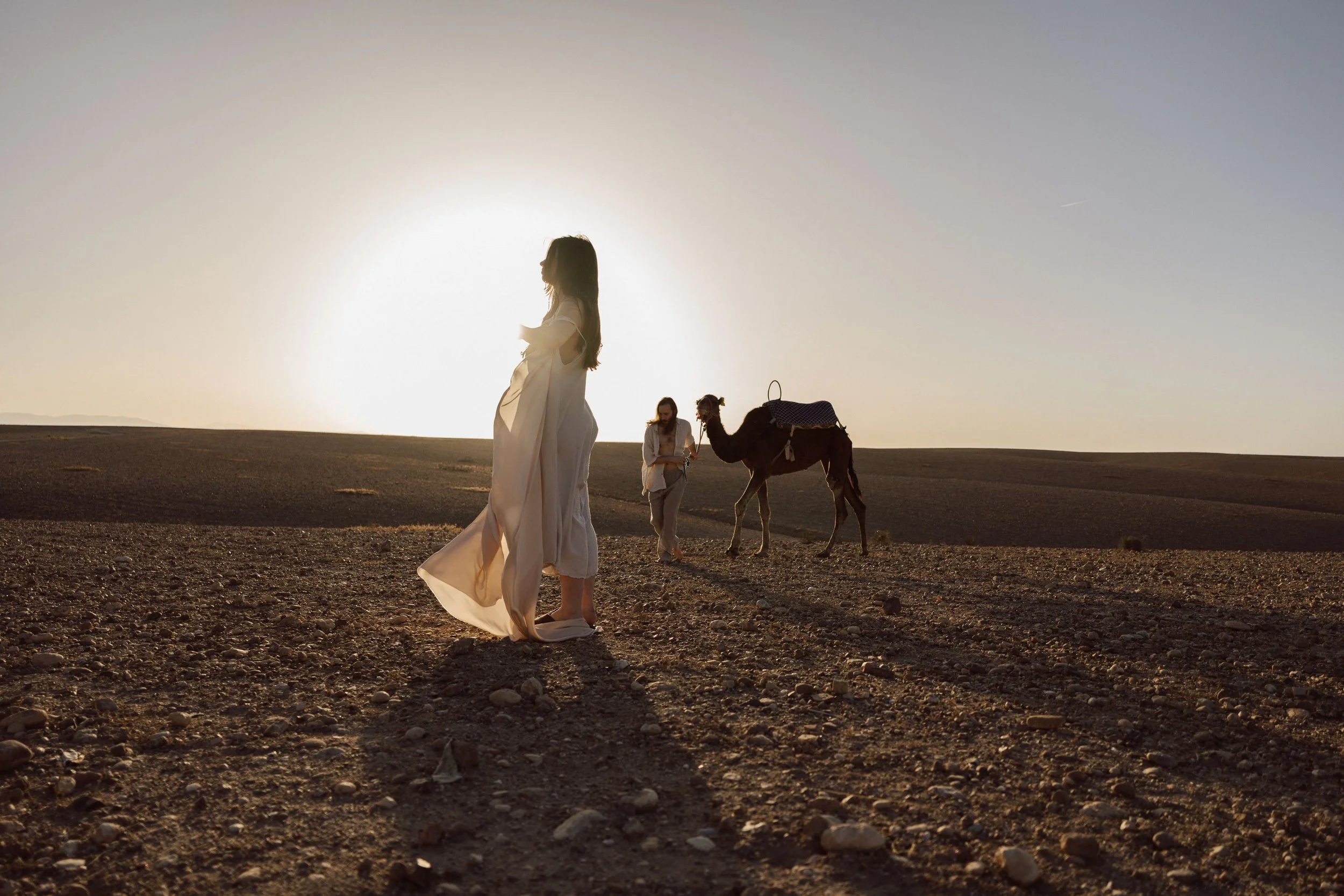 Séance elopement dans le désert marocain au coucher du soleil : une scène poétique entre couple, lumière dorée et chameau, capturée dans une atmosphère cinématographique par Nonante Émotions.