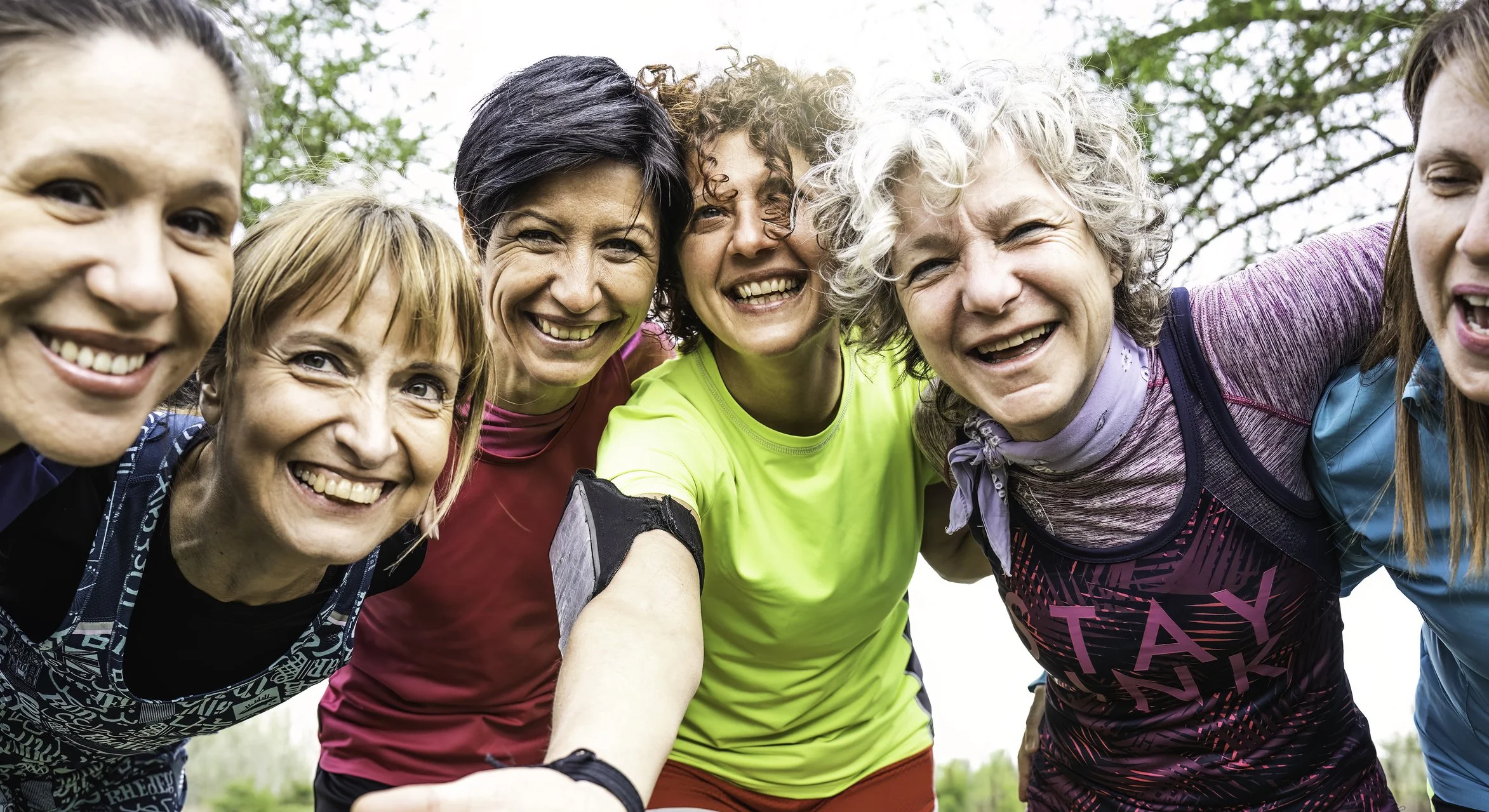 Group of smiling women and one man taking a selfie outdoors, surrounded by trees during daytime.