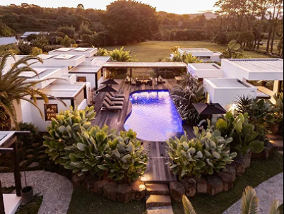 A luxurious outdoor pool area at sunset, surrounded by white modern buildings, lush greenery, and tropical plants, with seating areas and a scenic landscape in the background.
