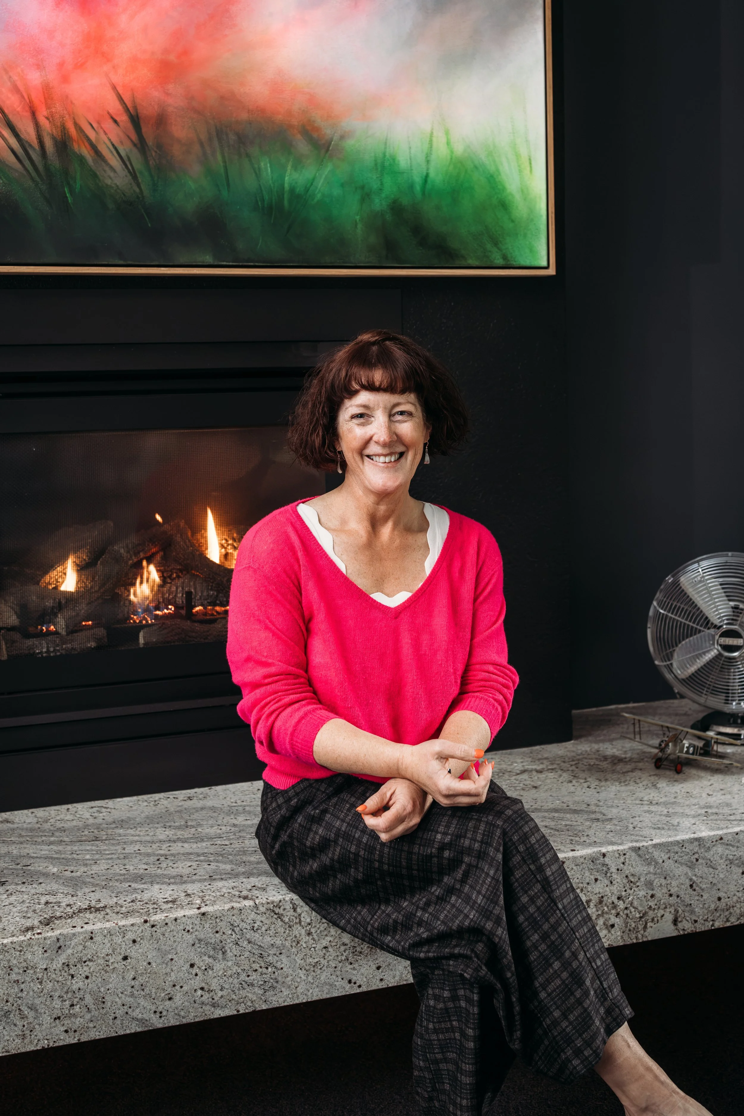 A woman with short brown hair sitting on a stone ledge in front of a fireplace, smiling, wearing a pink sweater and checkered pants, with colorful abstract art above the fireplace and a fan to her right.