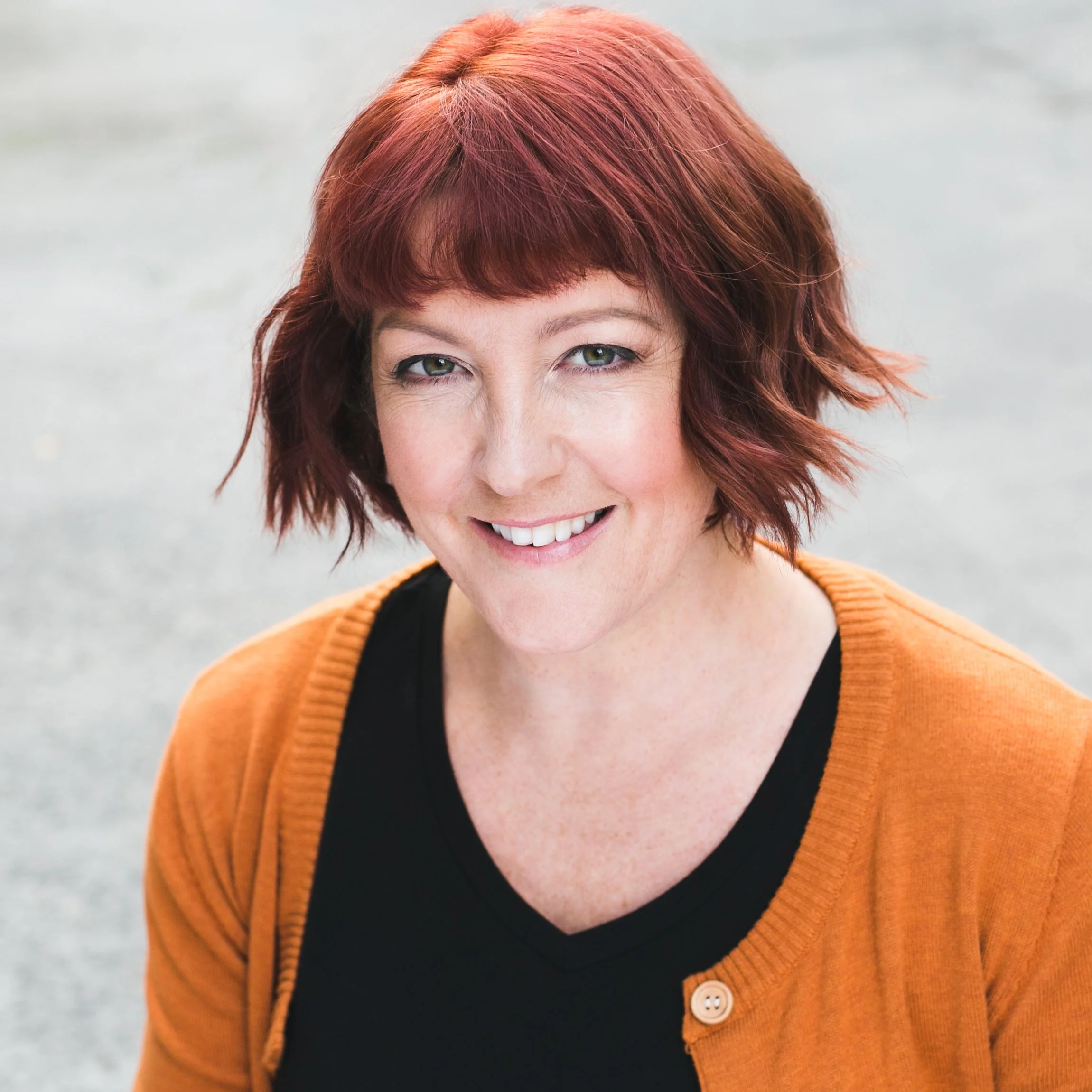 Close-up of a woman with short, wavy red hair, smiling and looking directly at the camera. She is wearing a black top and an orange cardigan with a button.