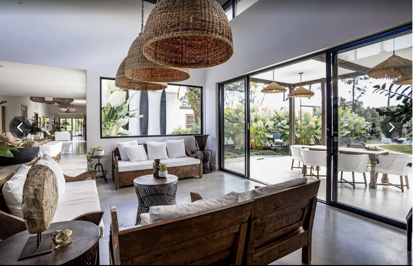 Modern living room with wooden furniture, white cushions, large glass sliding doors opening to an outdoor patio with greenery and outdoor seating, decorated with woven pendant lights.