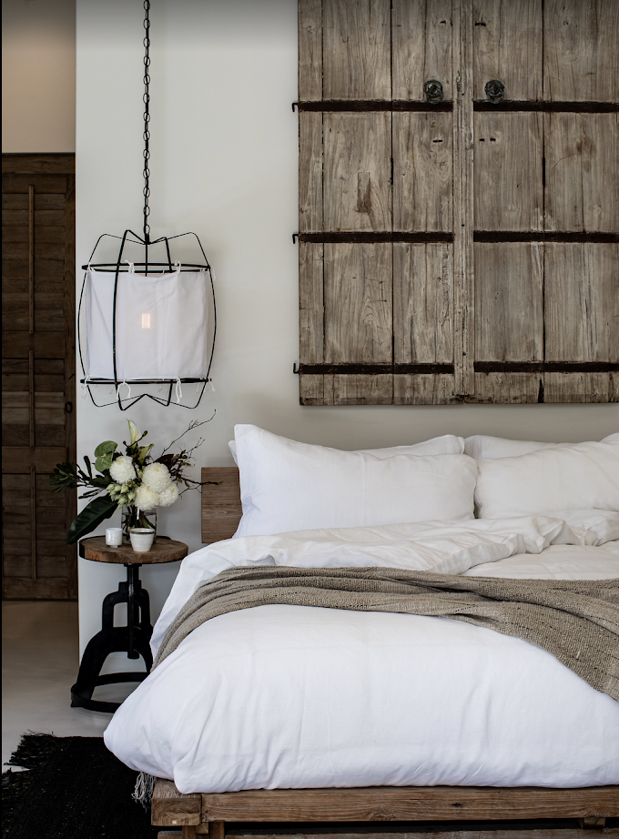A bedroom with a white bed, wooden headboard, and decorative wooden wall art, next to a small round wooden side table with a flower arrangement and a wall-mounted light fixture.