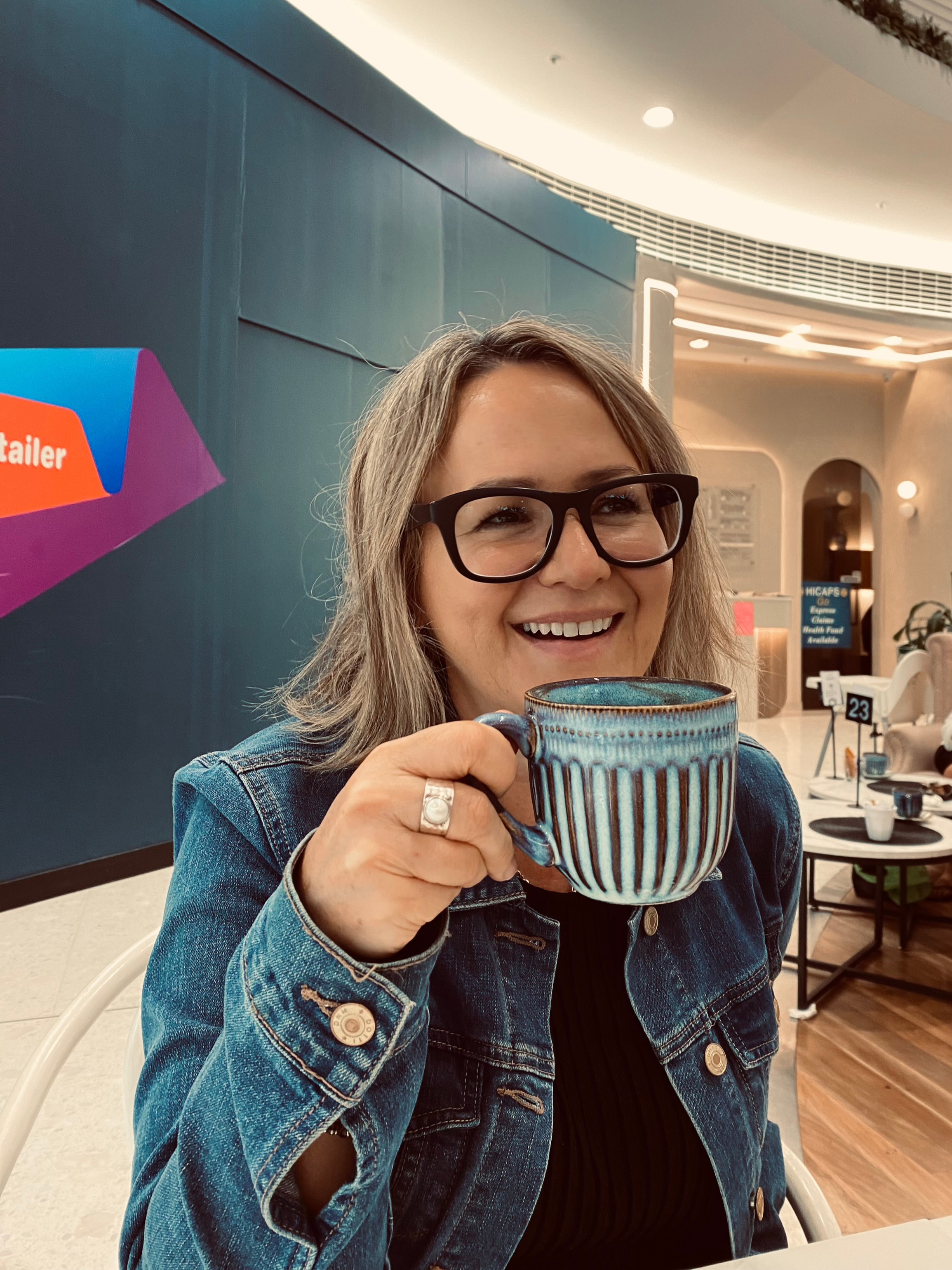 Woman with glasses smiling and holding a striped blue mug in a cafe.