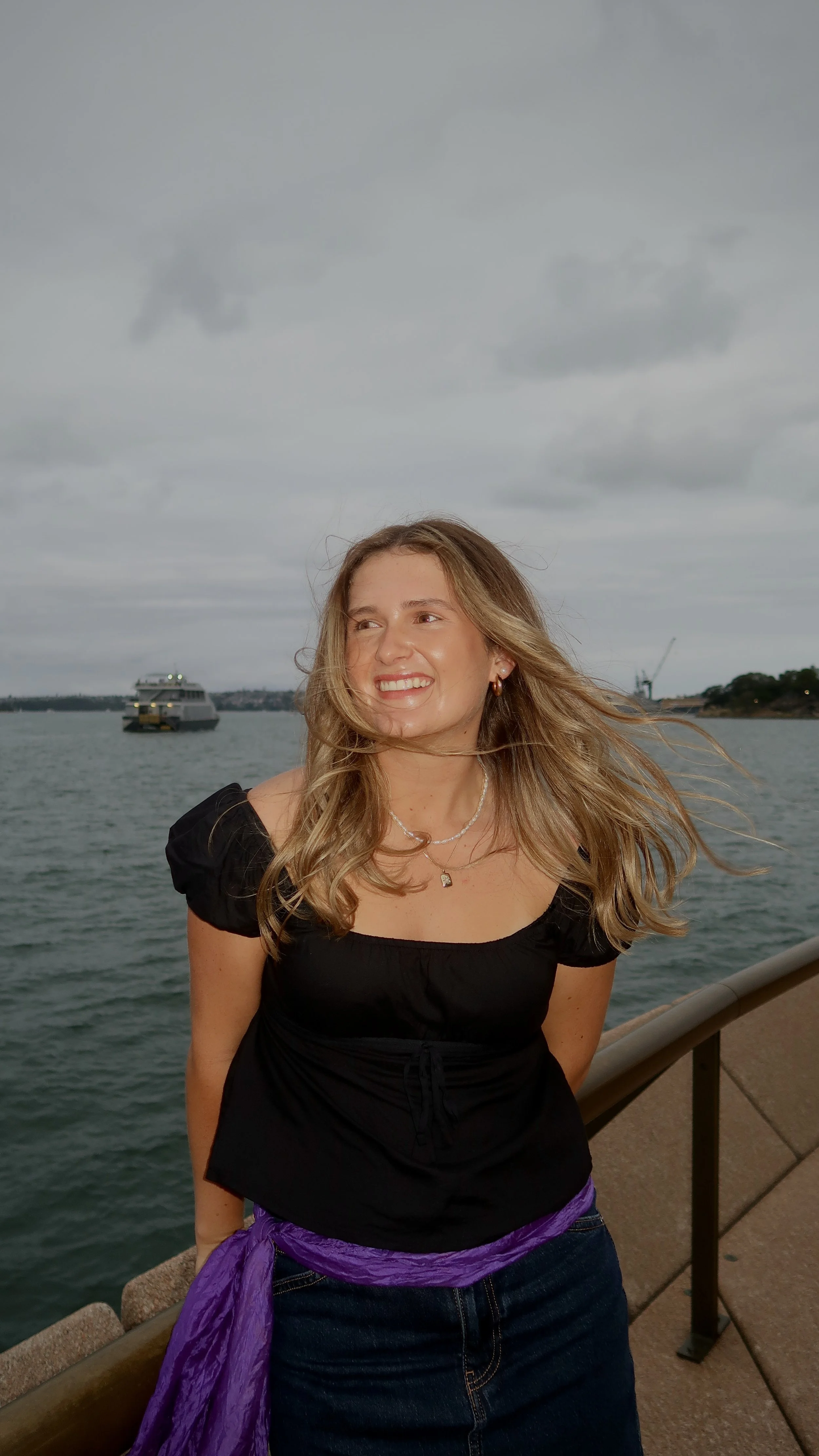 A young woman with wavy blonde hair stands by a waterfront on a cloudy day, smiling, with a boat and water in the background.