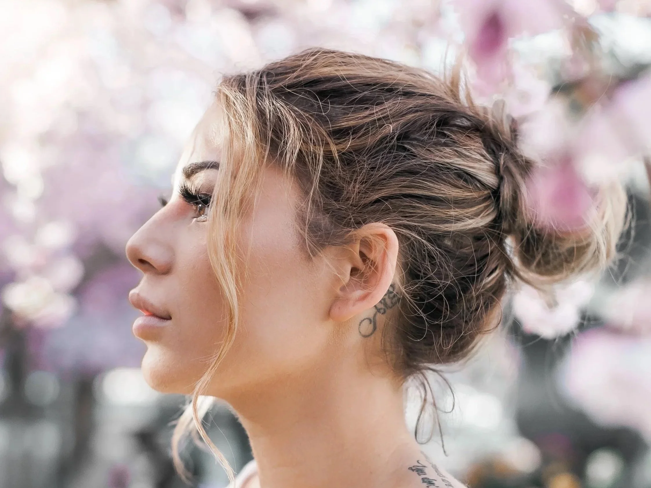 Side profile of a woman with light brown hair styled in an updo with some loose curls, standing in front of pink and purple blossoming trees.