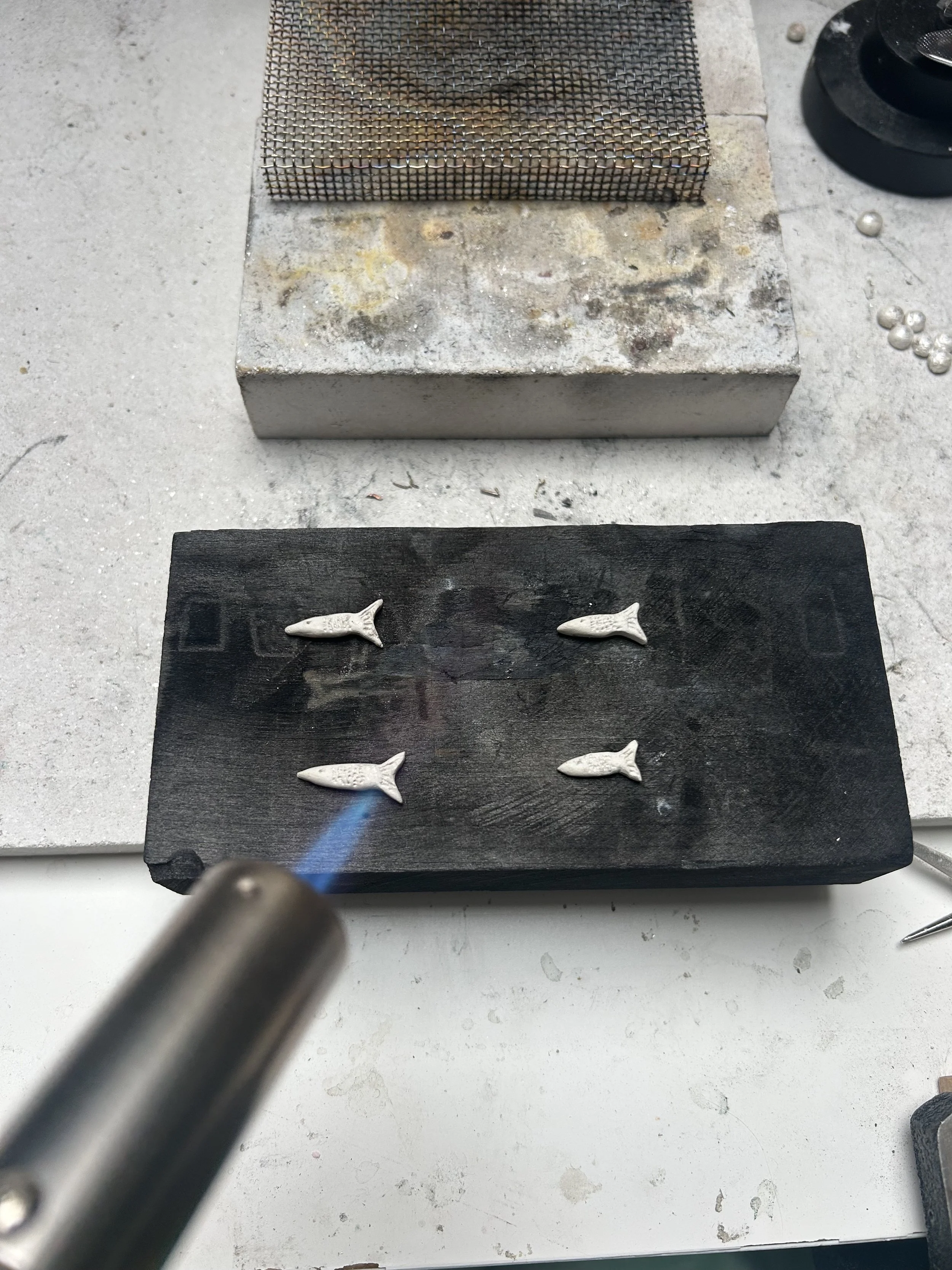 A small, silver-colored gas torch directing a blue flame at a black wooden surface with four white fish-shaped objects on it. Silver clay being turned into solid silver jewellery piecs