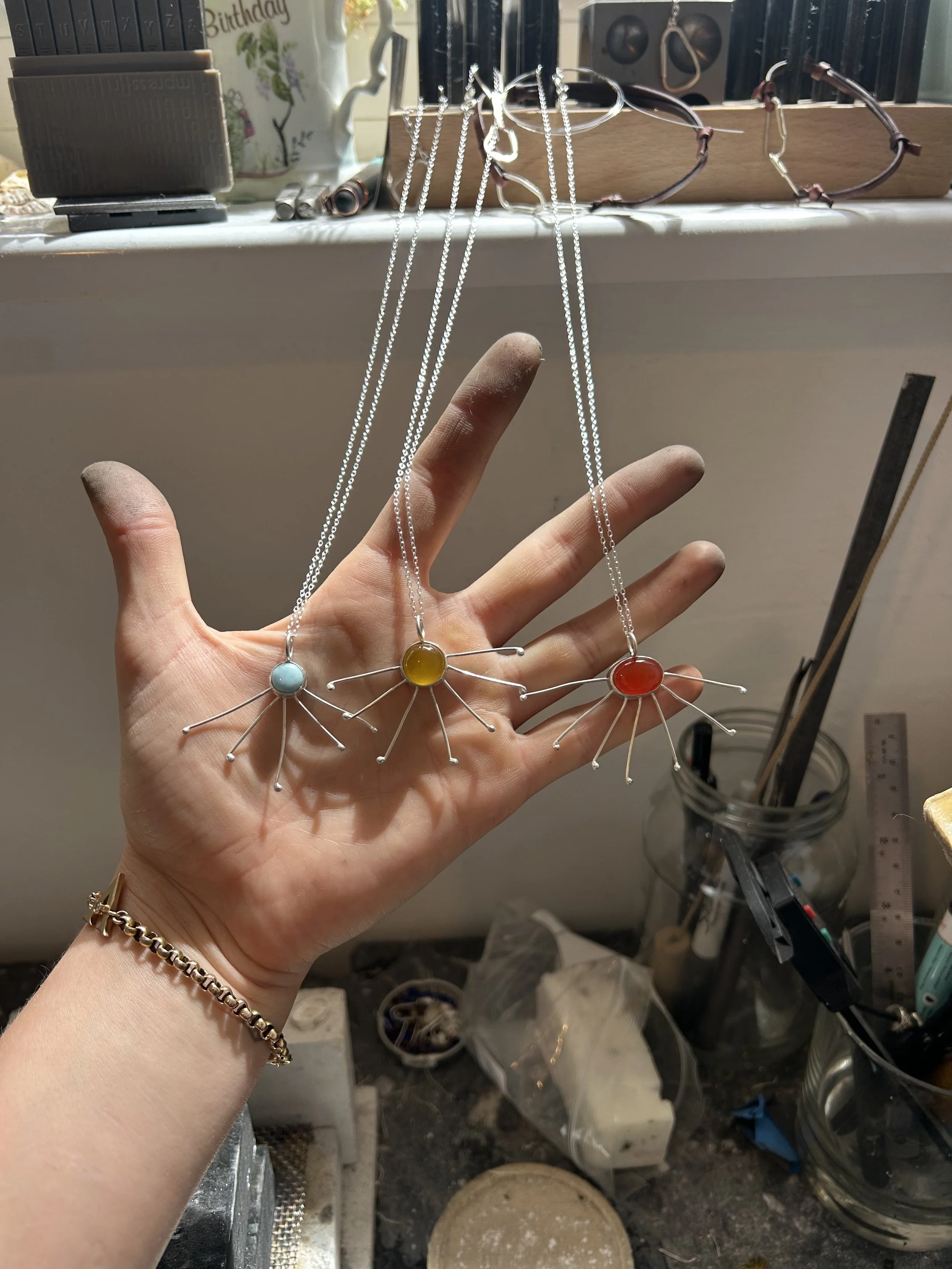 Hand holding three necklaces with colorful circular pendants: blue, yellow, and red, in a jewelry workspace with tools, books, and miscellaneous items in the background.