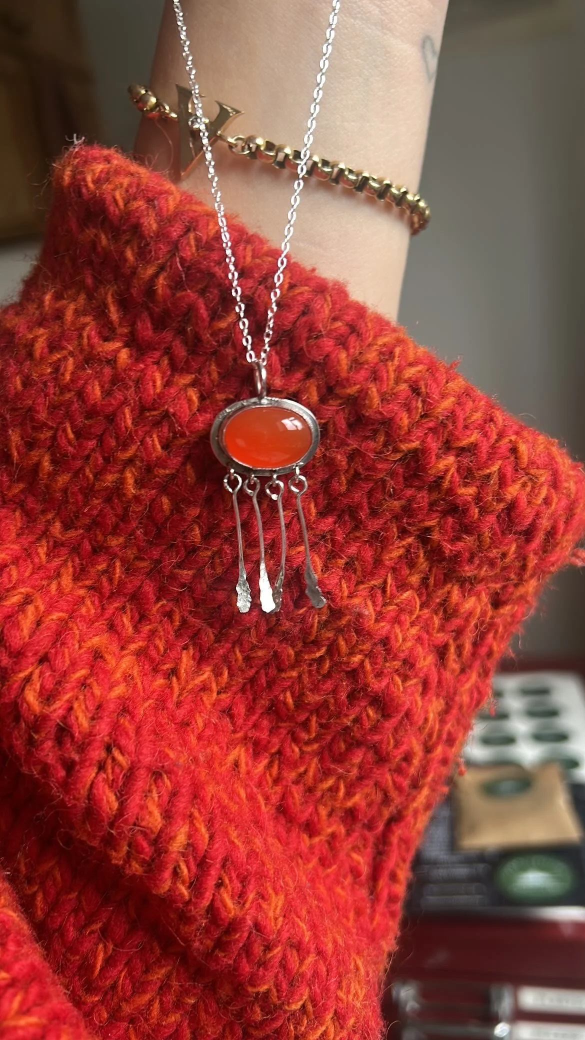 Close-up of a person's arm wearing a red knitted sweater. A silver necklace with an orange pendant, and sterling silver adornments. 