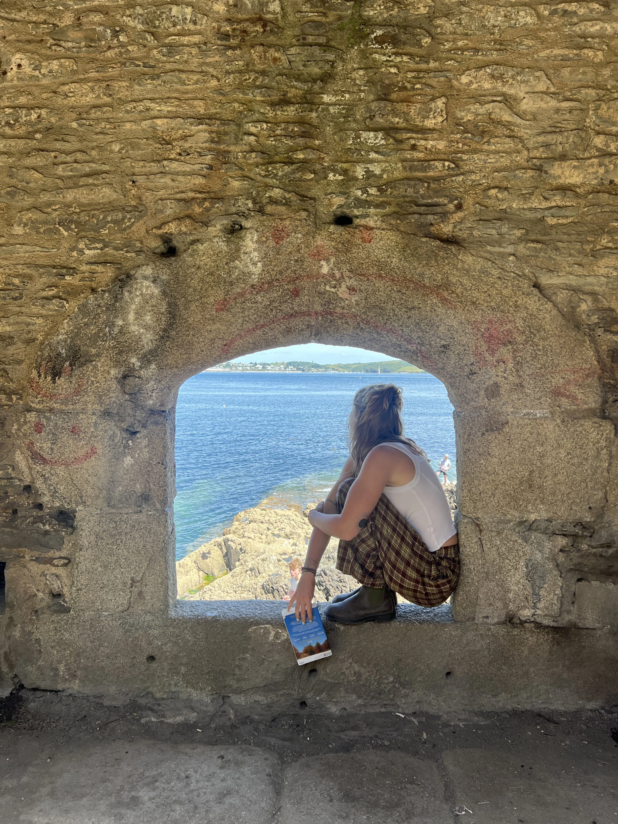 Person sitting in a small stone window, overlooking water and distant shoreline with green hills, at a historic site.