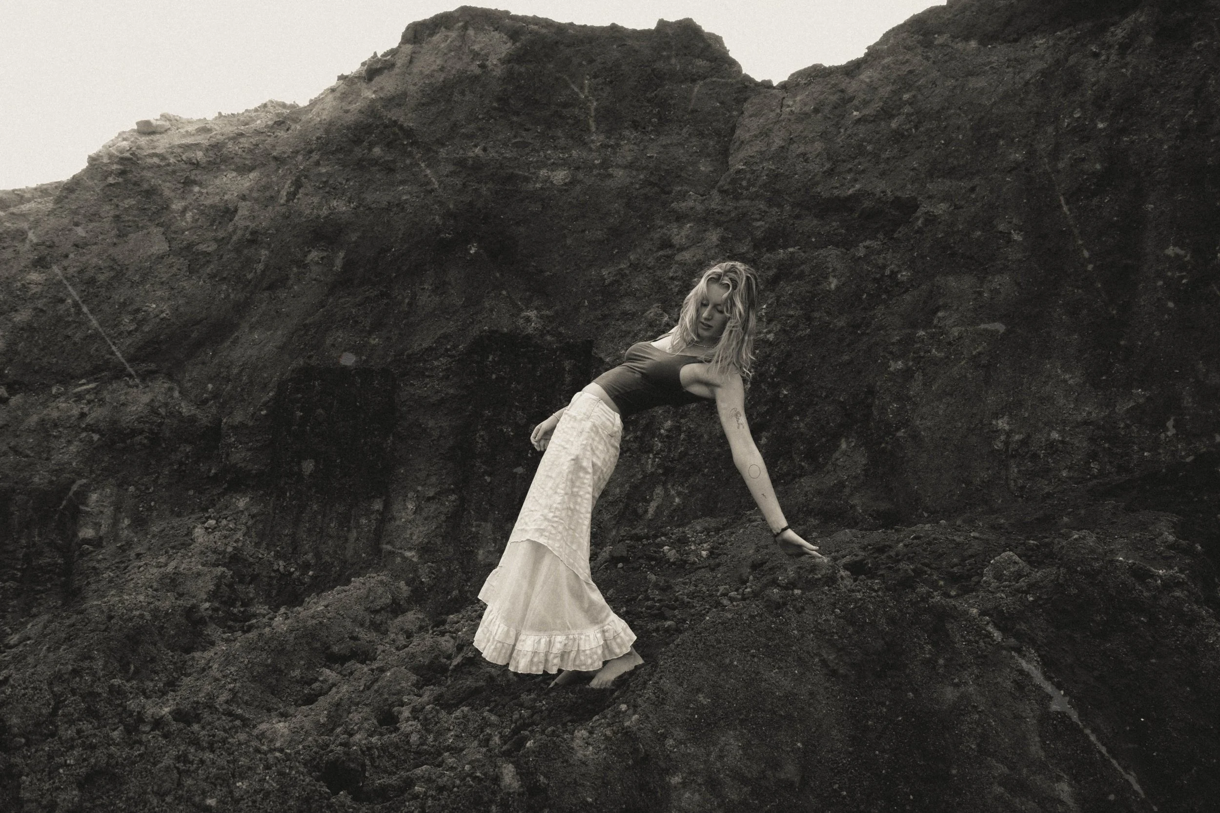 A woman with long blonde hair, wearing a dark sleeveless top and a long white skirt, is leaning back with one hand on the ground and her head tilted downward, standing in front of a large dark rocky mound.