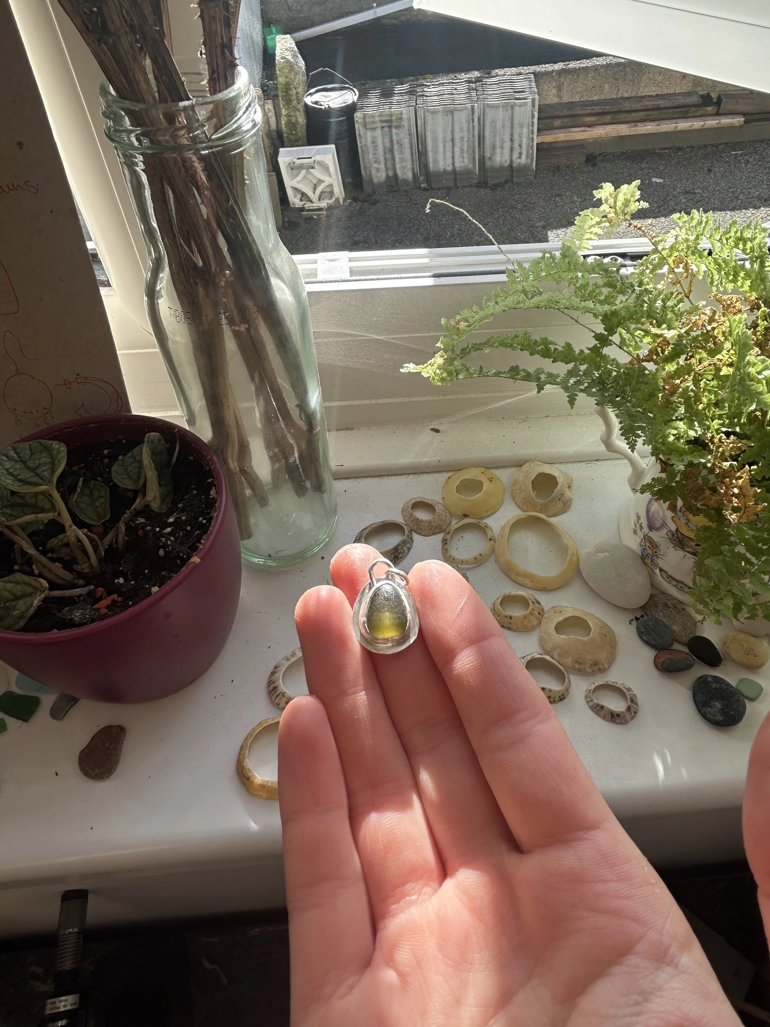 Hand holding a ring with a yellow-green stone over a collection of rings and stones on a windowsill, with plants and outdoor objects visible through the window.