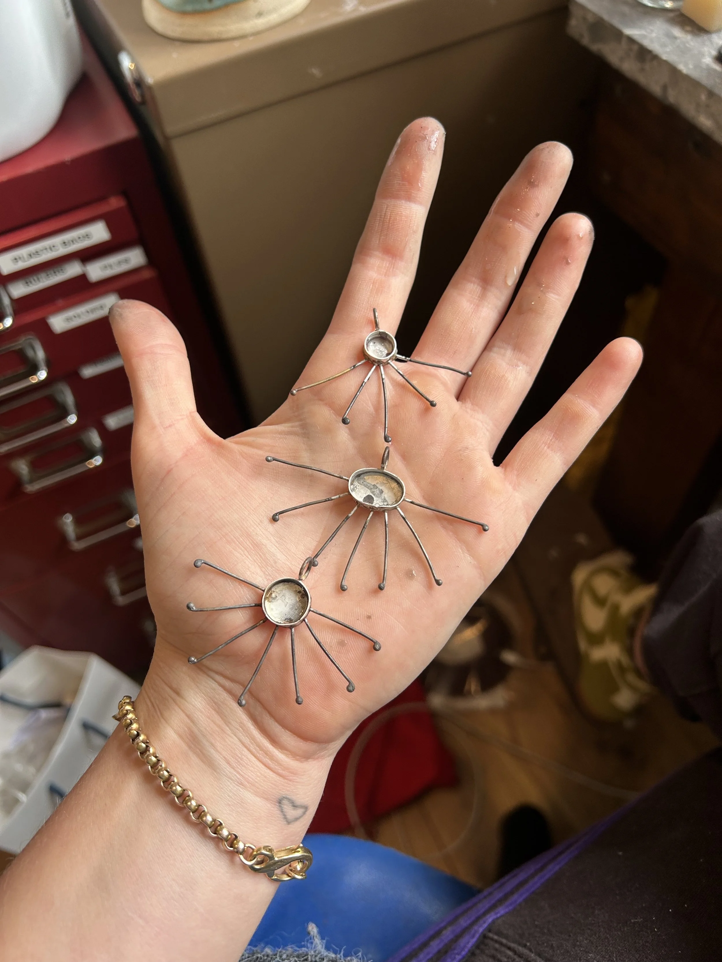 A person's hand, holding unfinished sterling silver sun shape pendants