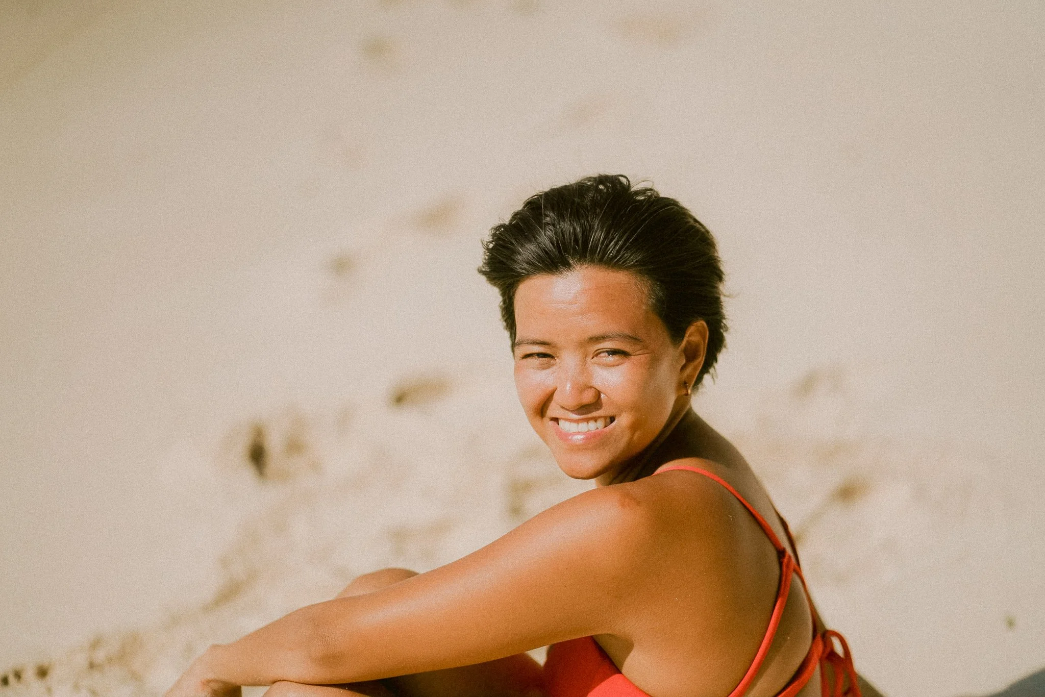A woman with short black hair, wearing a red top, smiling and squinting outdoors against a blurred background.