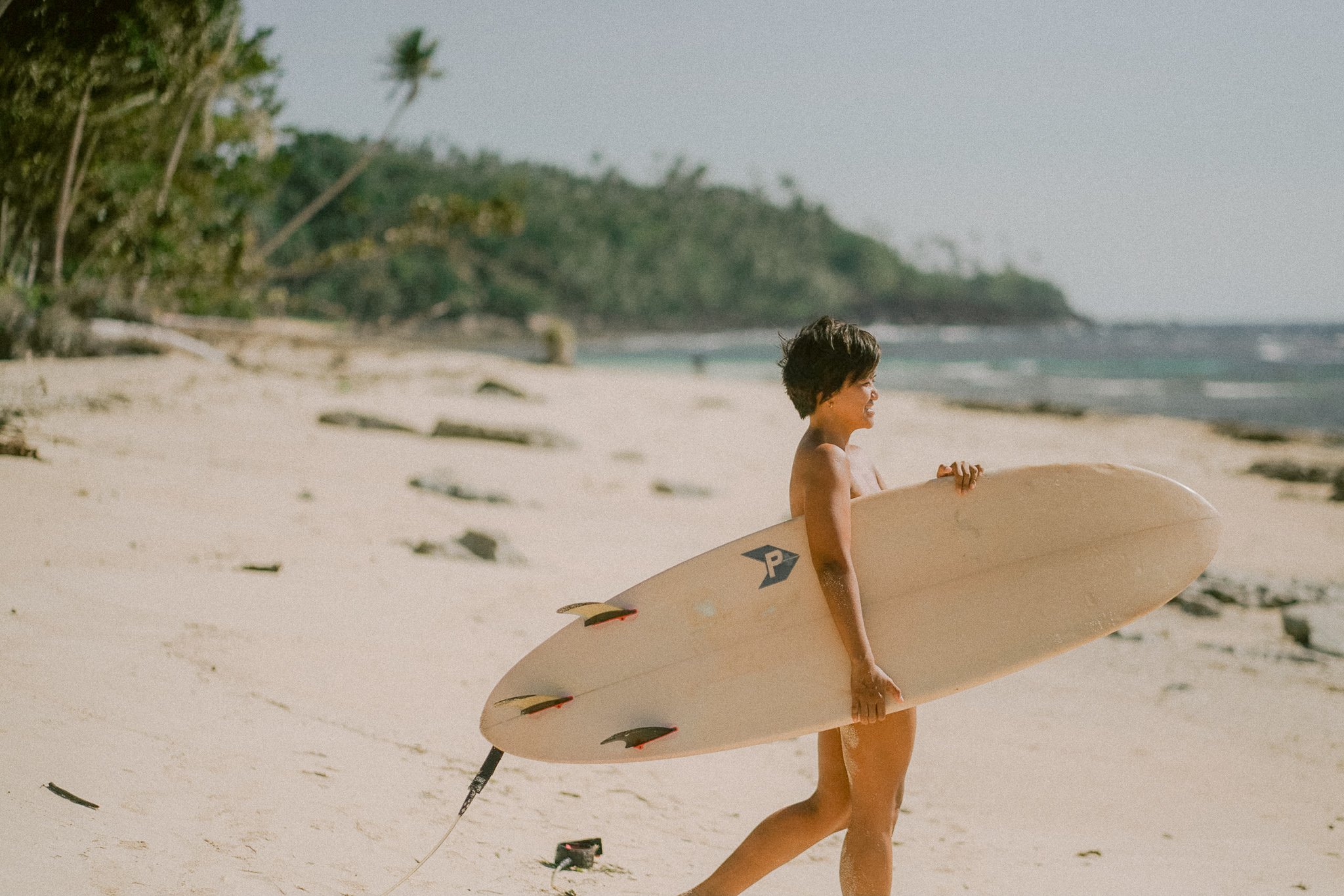 A young boy with short dark hair holding a surfboard on a sandy beach, with greenery and a hill in the background, under a cloudy sky.