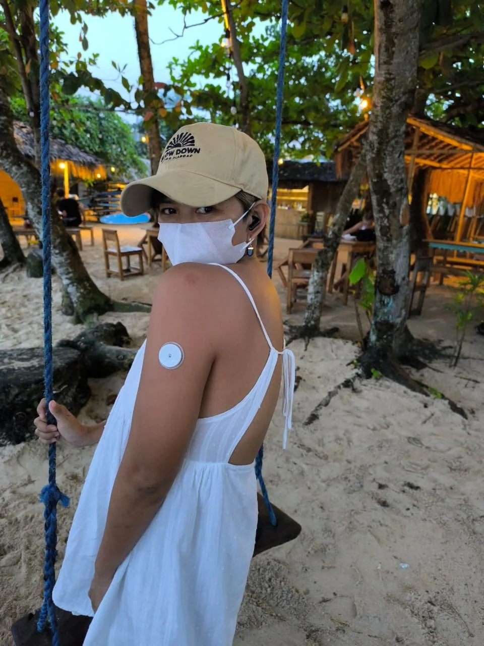 A woman wearing a white dress, beige cap, and face mask sitting on a swing at a beachside area with trees, sand, and outdoor seating in the background. She is wearing a CGM (Continuous glucose Monitoring) for her diabetes.