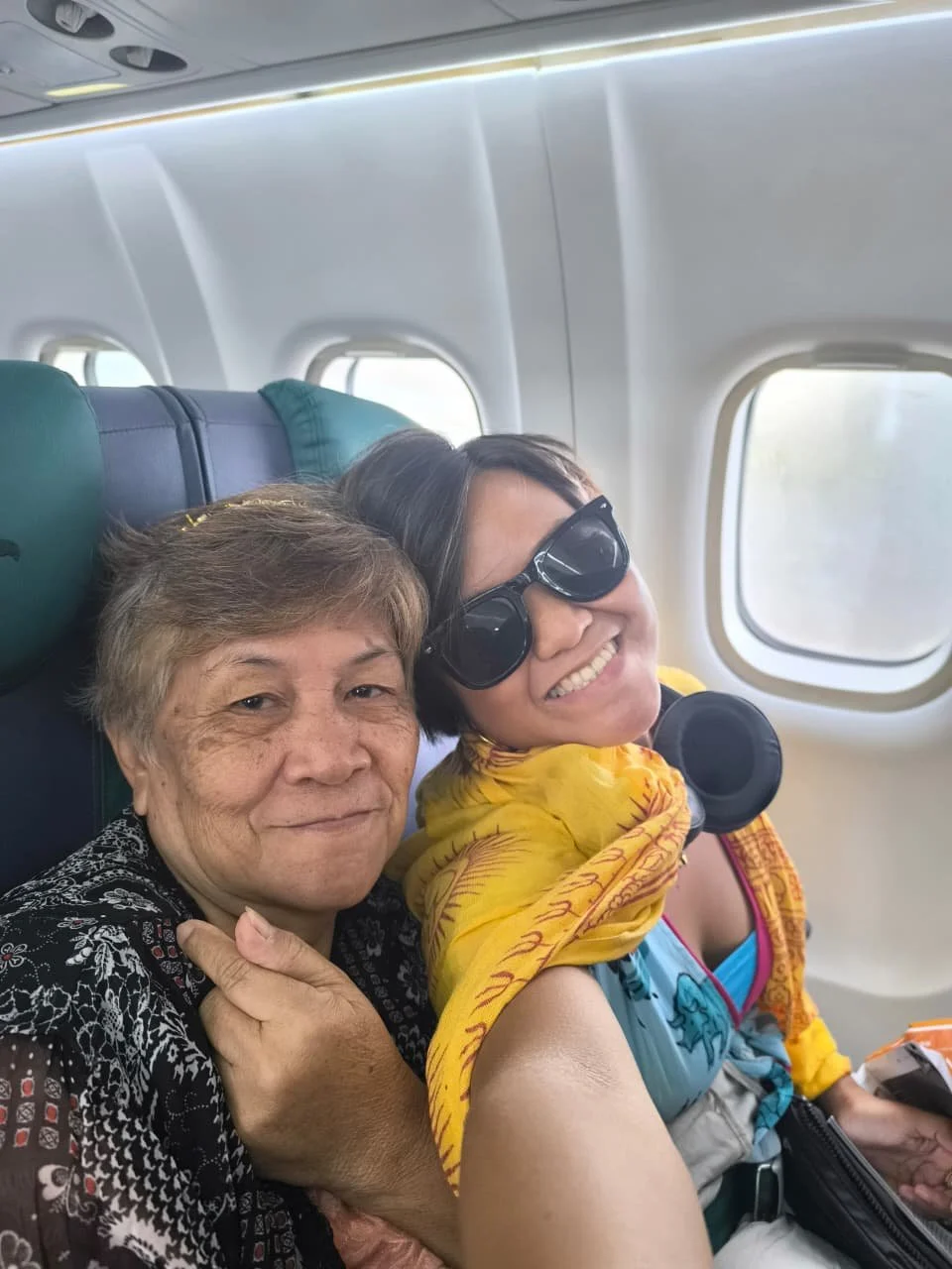 Two women sitting close together on an airplane, smiling at the camera. One woman has short hair, is wearing a black patterned shirt, and has her hand on the other woman's shoulder. The other woman is wearing sunglasses and a colorful jacket, and is holding a phone.