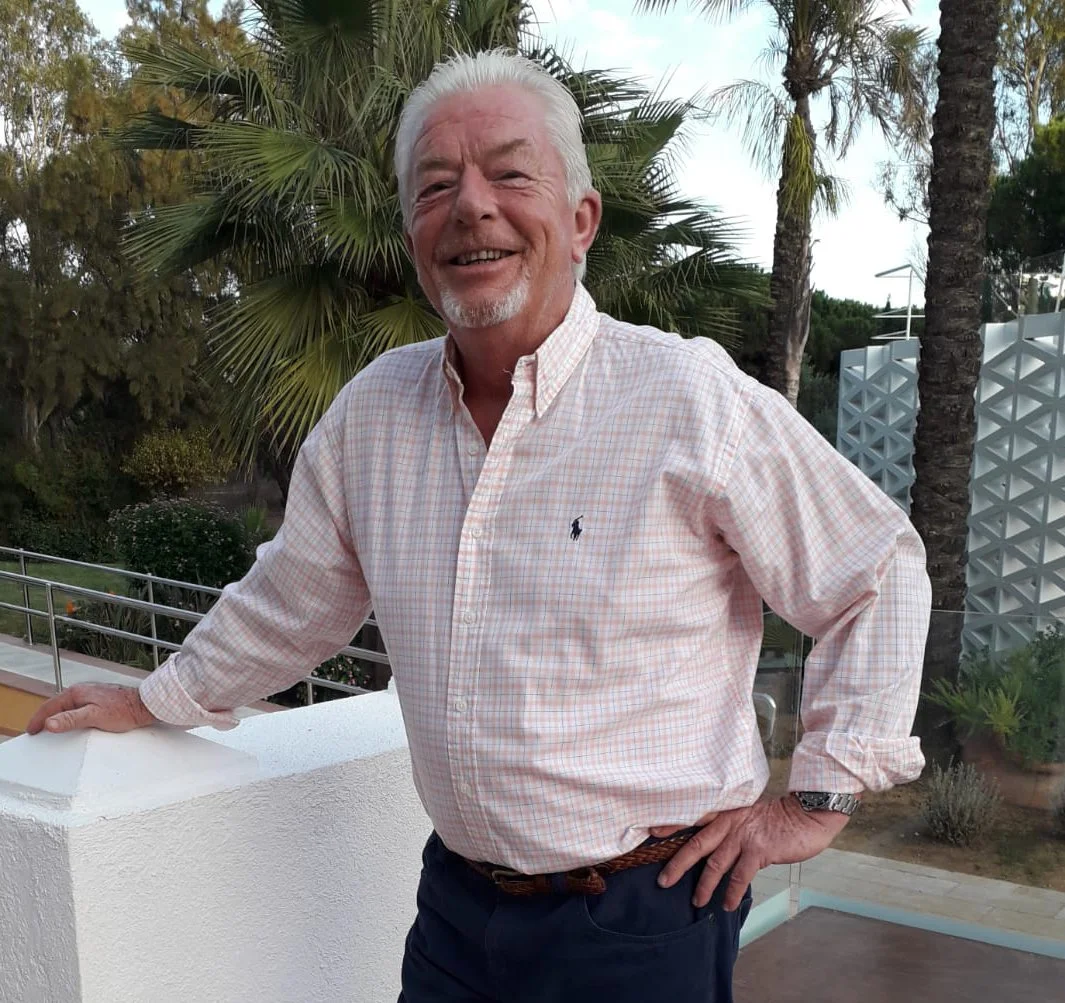 Older man with white hair and beard smiling, wearing a light-colored checkered shirt and dark pants, standing outdoors with a background of palm trees and greenery.