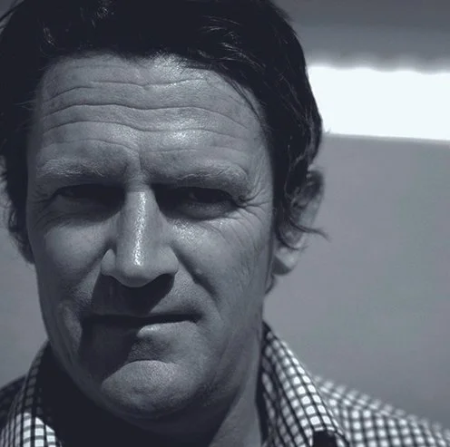 Close-up black and white portrait of a man with dark hair, prominent facial features, and wearing a checkered shirt.