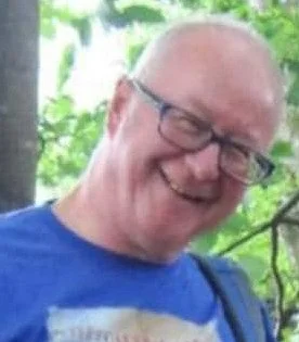 A man with glasses and a blue shirt smiling outdoors with greenery in the background.