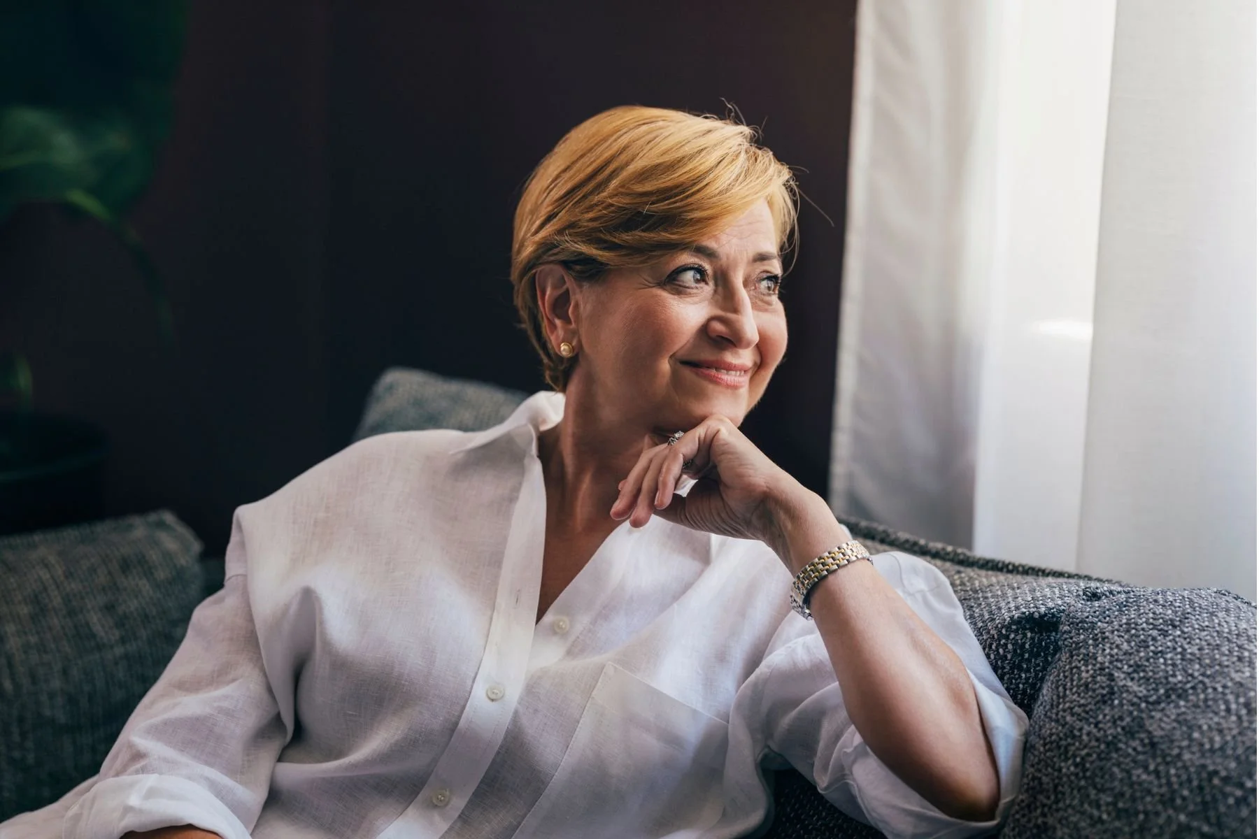A woman with short blond hair, wearing a white button-up shirt and a gold watch, sitting on a gray couch near a window with white curtains, smiling thoughtfully.