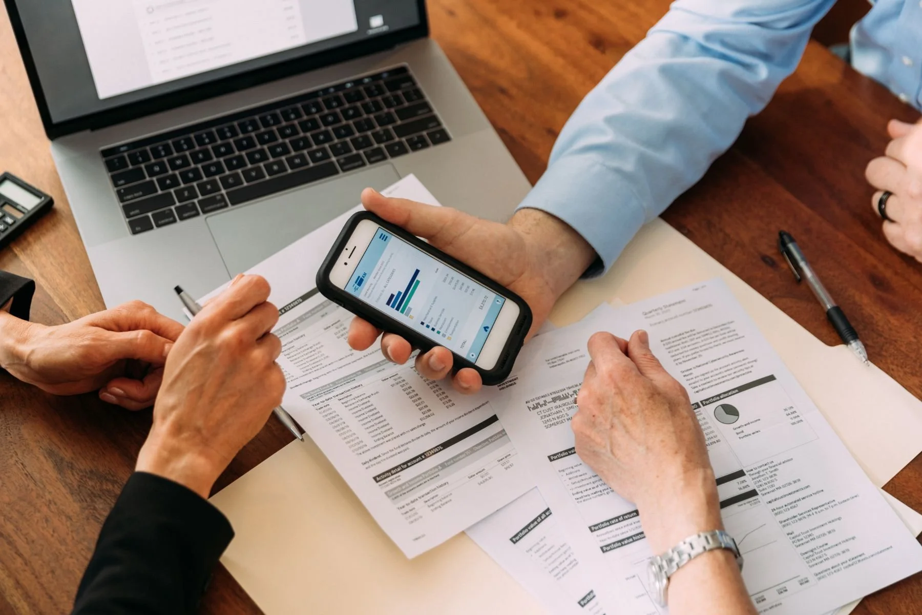 Two people reviewing financial documents with a laptop, smartphone displaying a bar chart, and pens on a wooden desk.