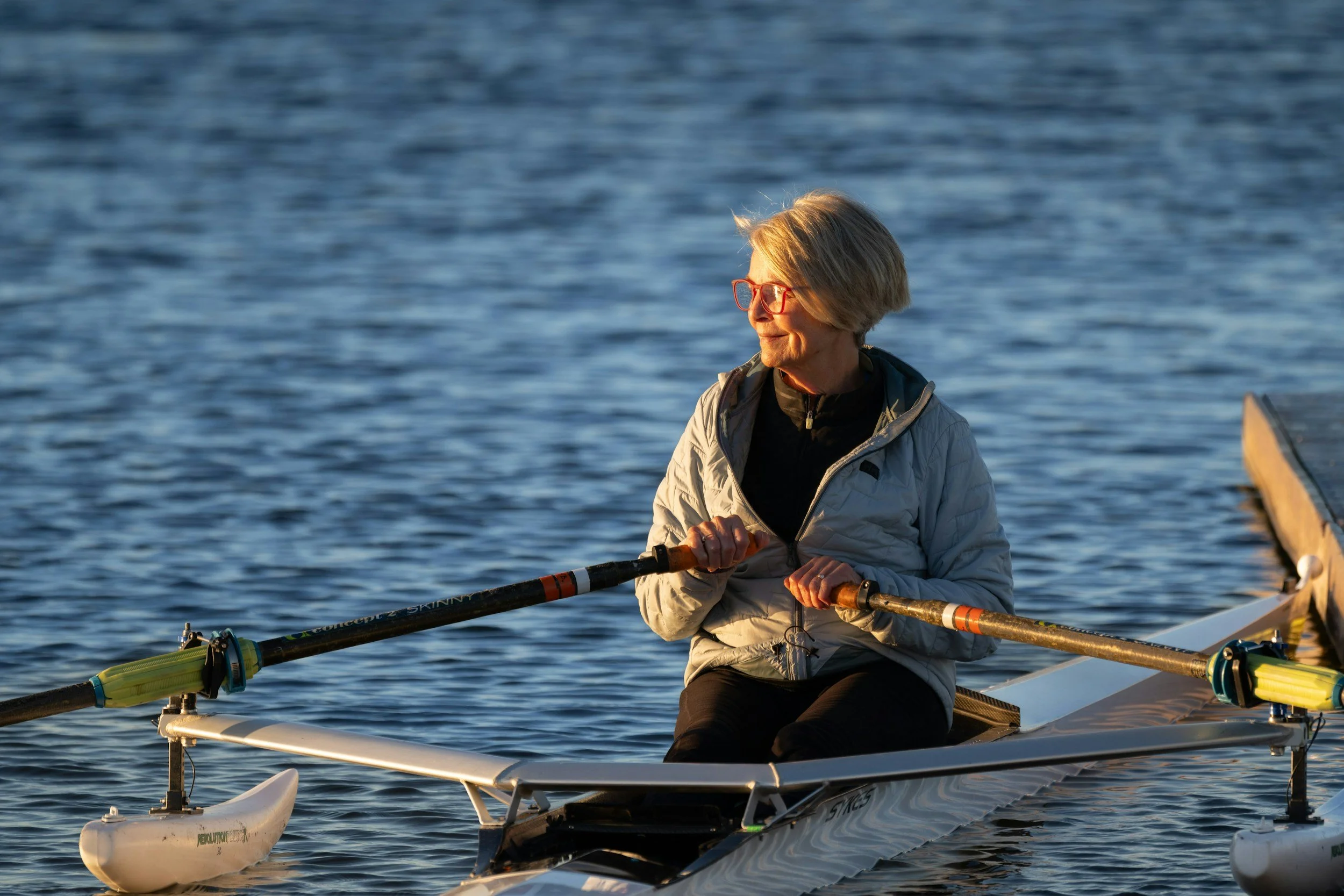 A woman with short blonde hair and red glasses rowing a small boat on a calm body of water during sunset.