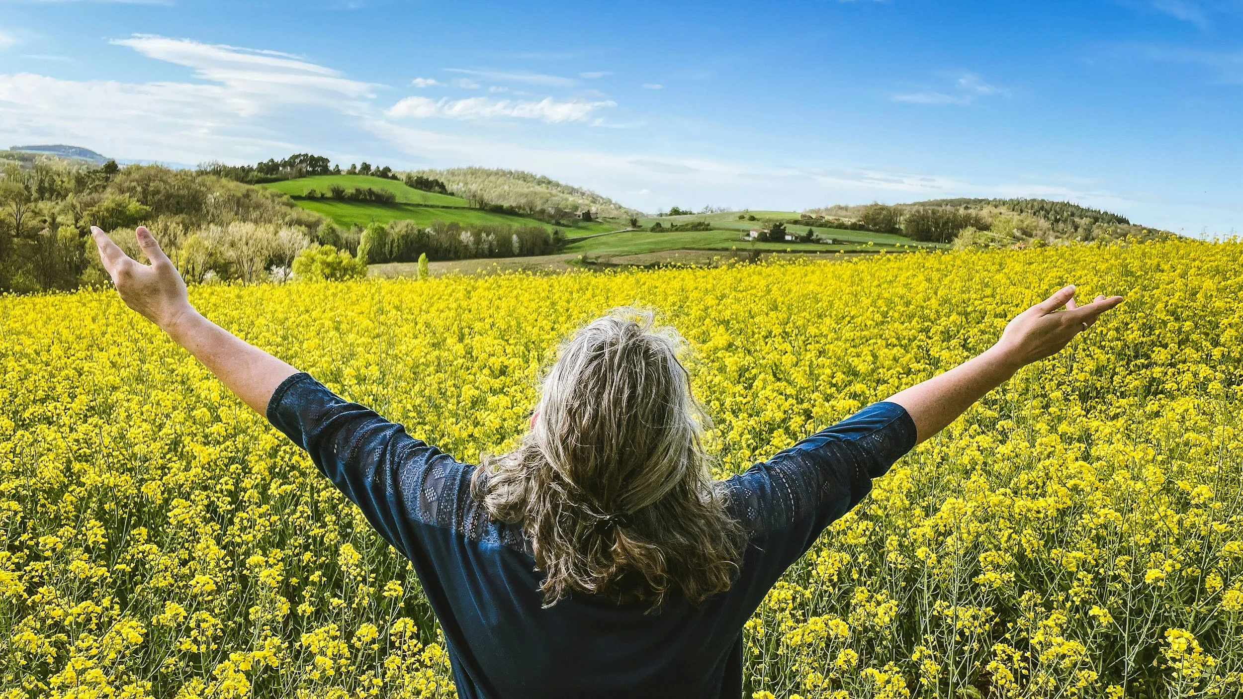 Person with gray hair standing in a field of yellow flowers with arms raised, overlooking rolling green hills and a partly cloudy blue sky.