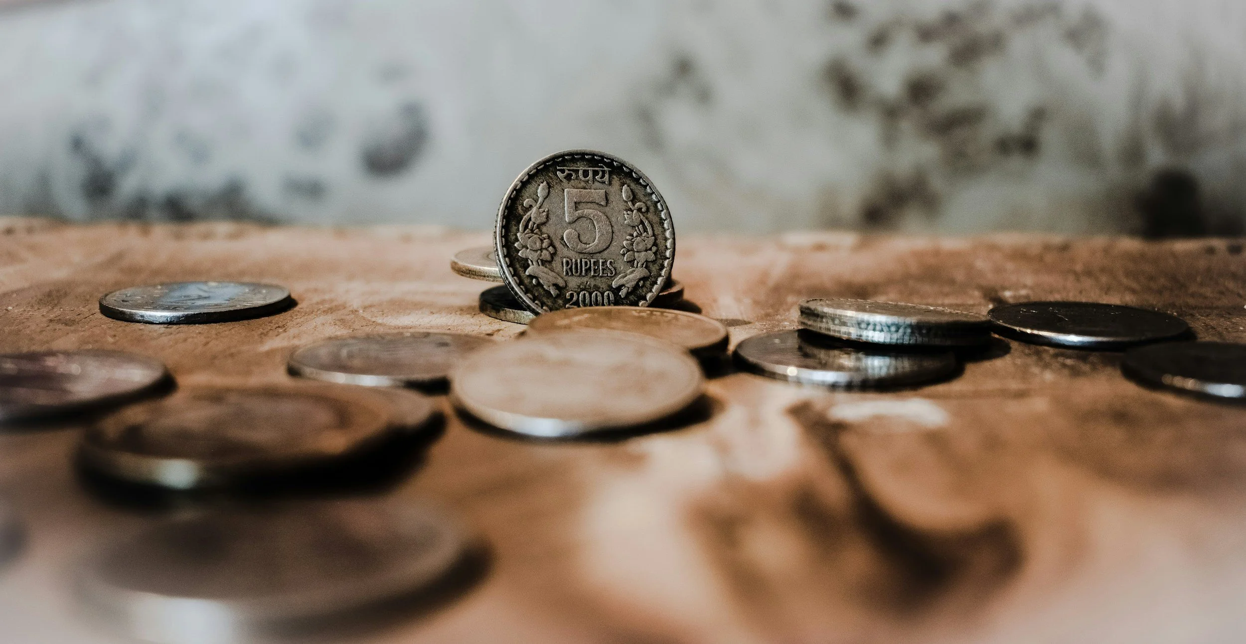 Close-up photograph of scattered Indian coins on a textured surface, with a focus on a 5 Rupees coin in the center.