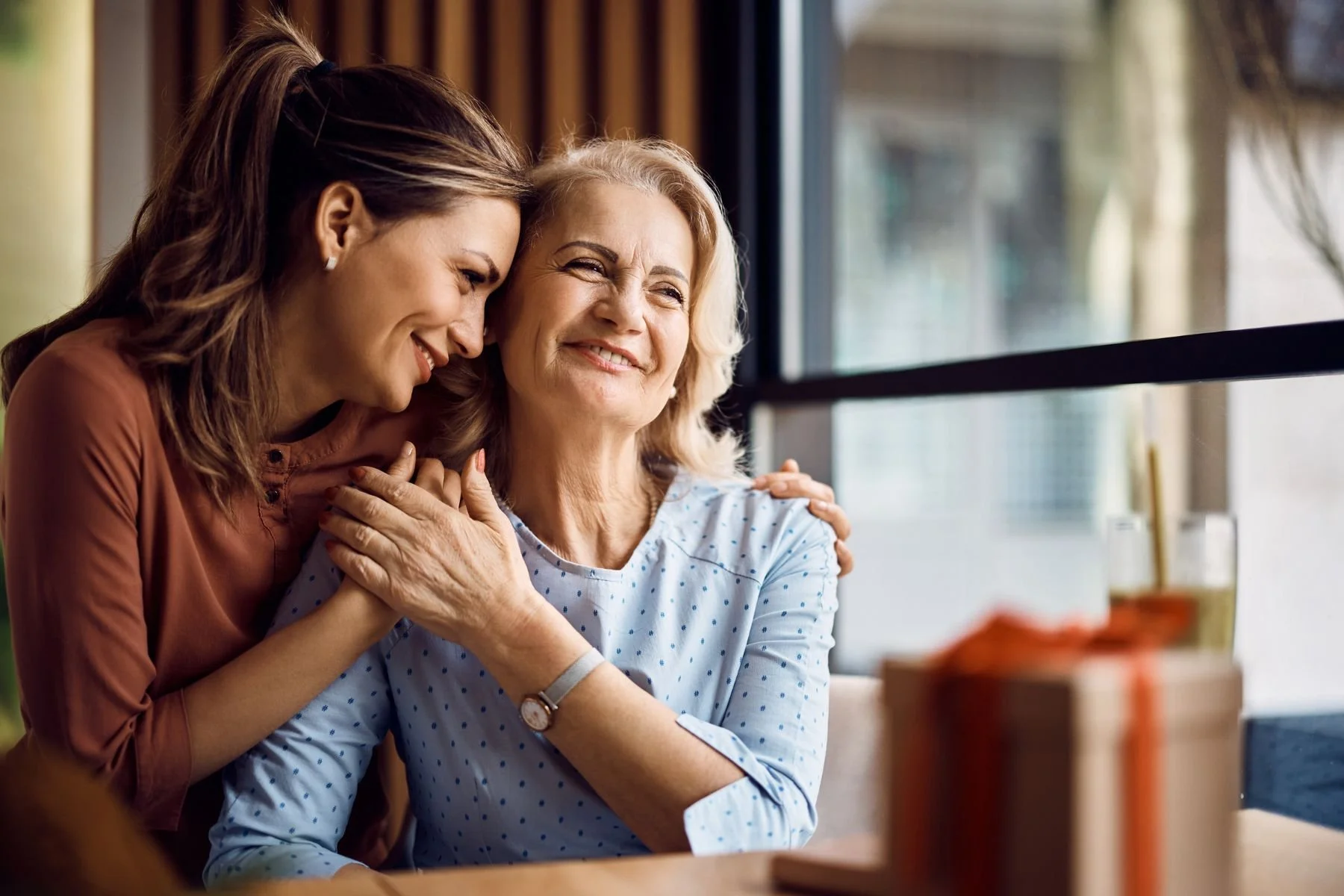 A young woman comforting an older woman, both smiling and sitting close together near a window.