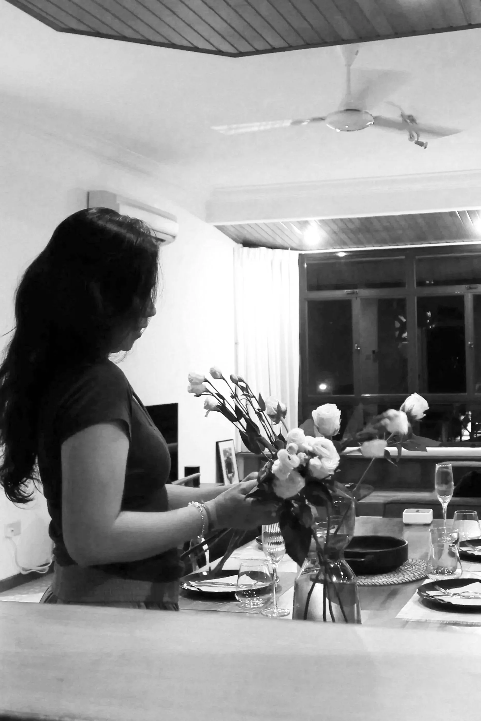 A woman with long dark hair holding a bouquet of flowers in a dining room. There are glasses, plates, and a vase with flowers on the table. The room has a wooden ceiling and a window.