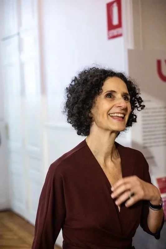 Mujer con cabello rizado, vestido marrón, sonriendo en un entorno interior con paredes blancas y señal de emergencia roja.