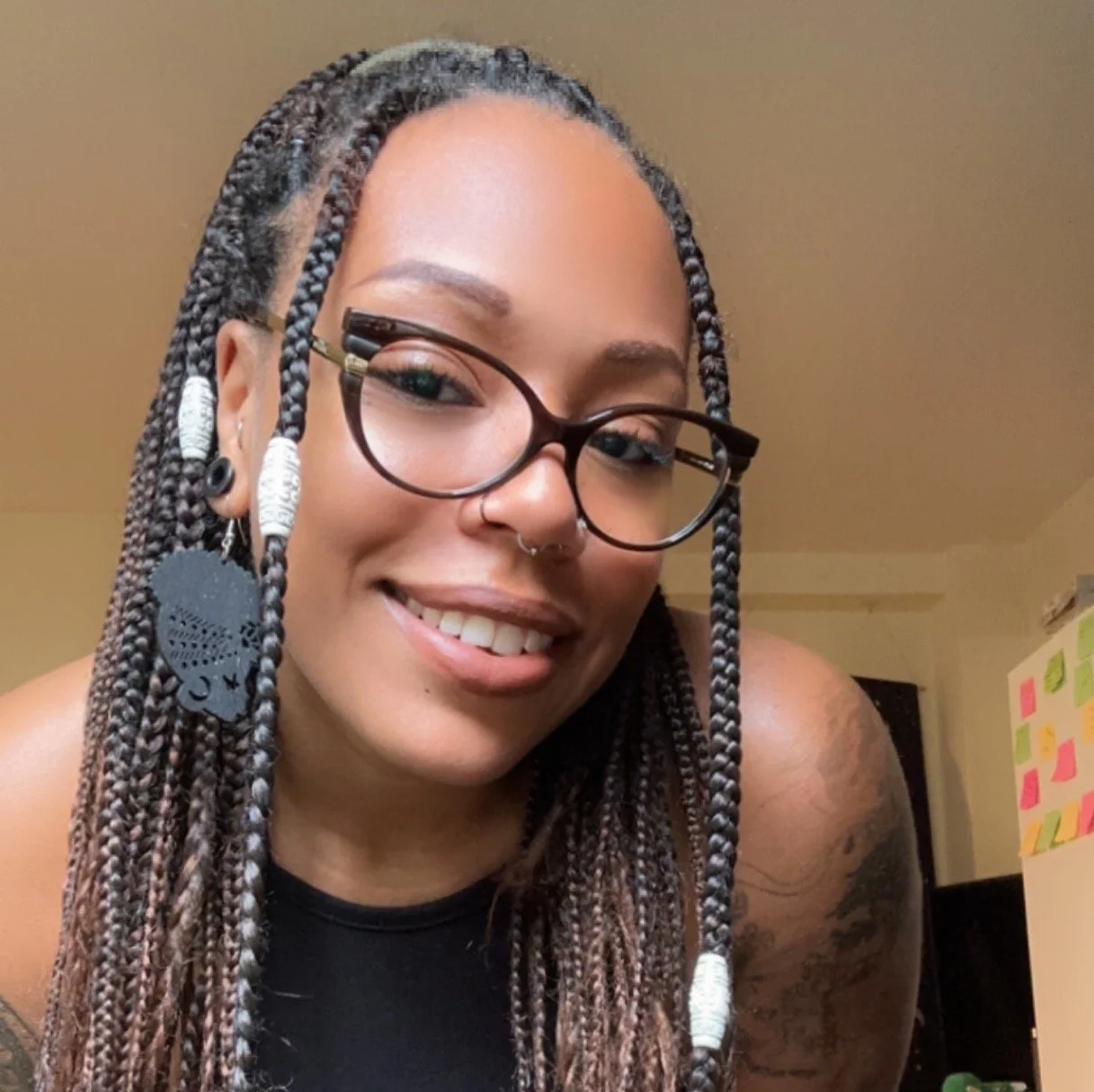 Picture of TJ Williams; a Black mixed-race person with braids, big earrings, and glasses,  smiling at the camera.