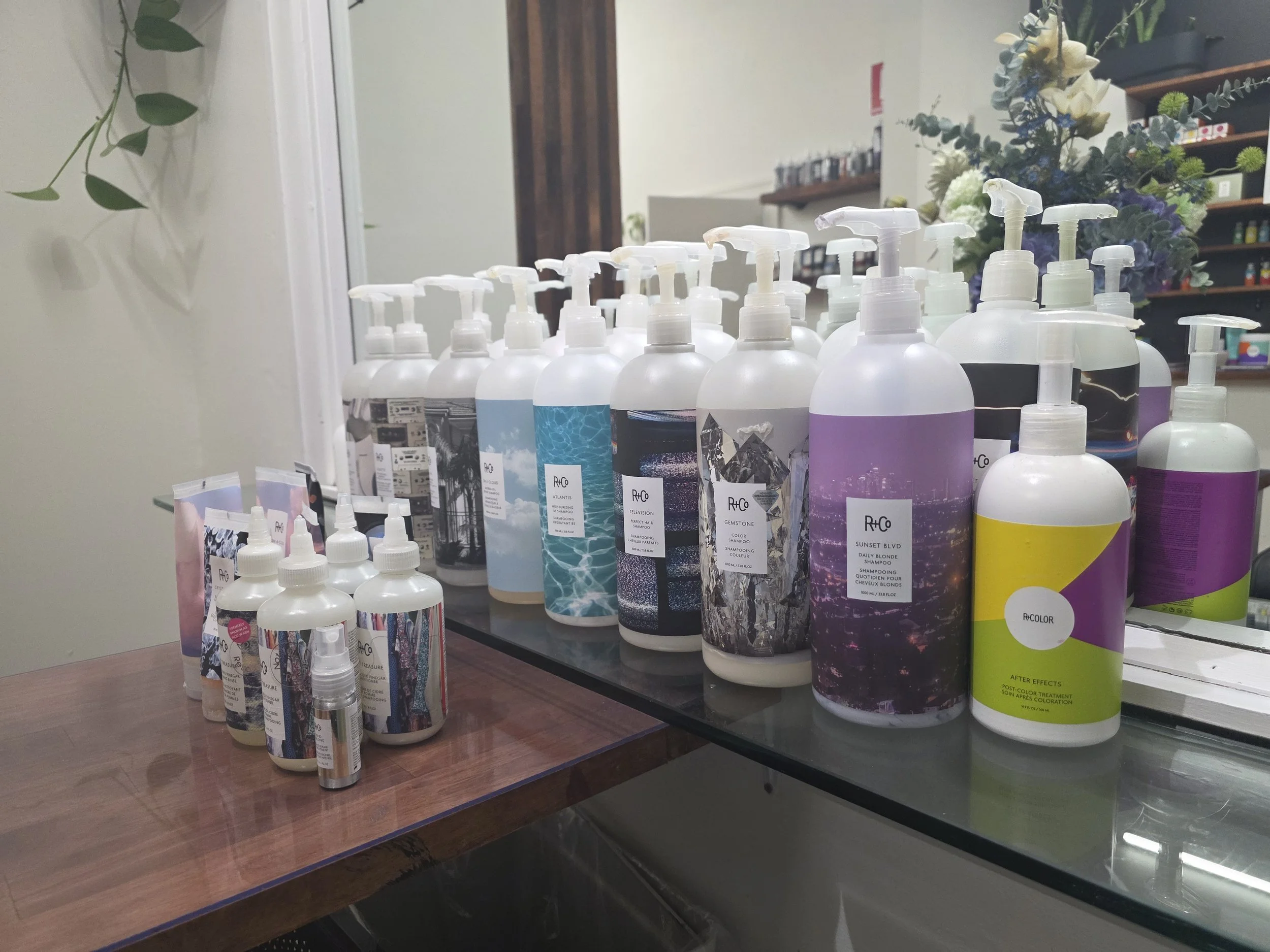 Display of various bottles of hair and skin care products on a table in a salon or store, with some flowers and plants in the background.
