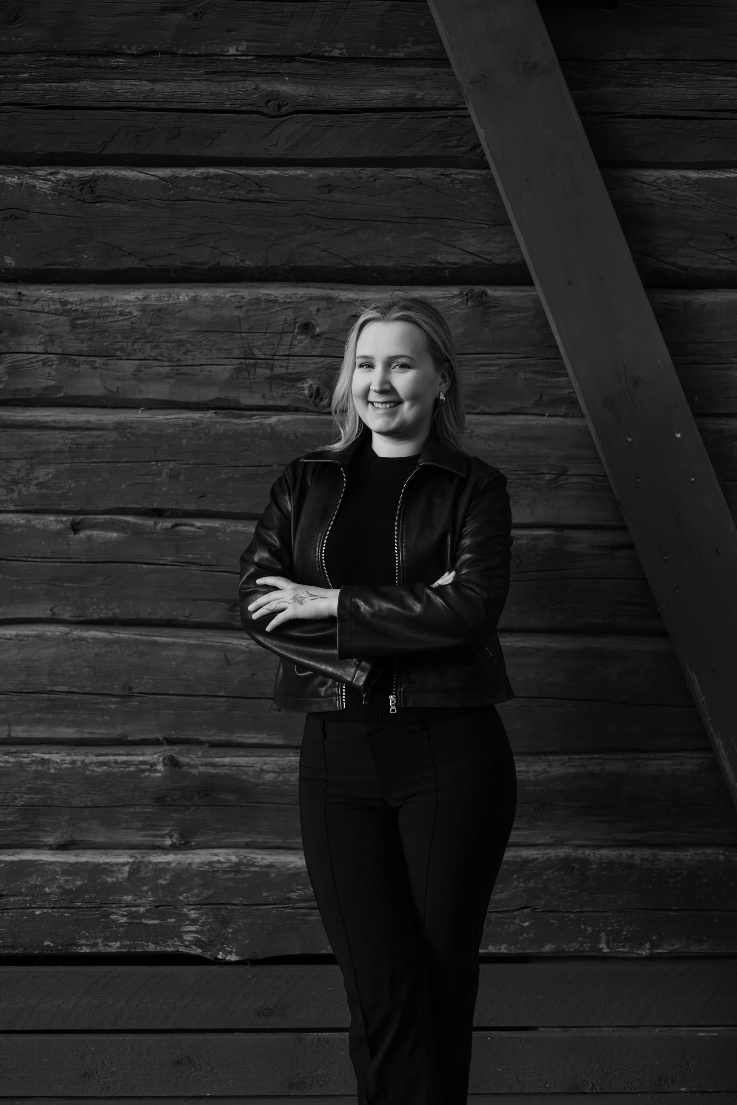 A woman with shoulder-length hair, smiling, wearing a leather jacket and black pants, standing with her arms crossed in front of a wooden wall with diagonal beams.