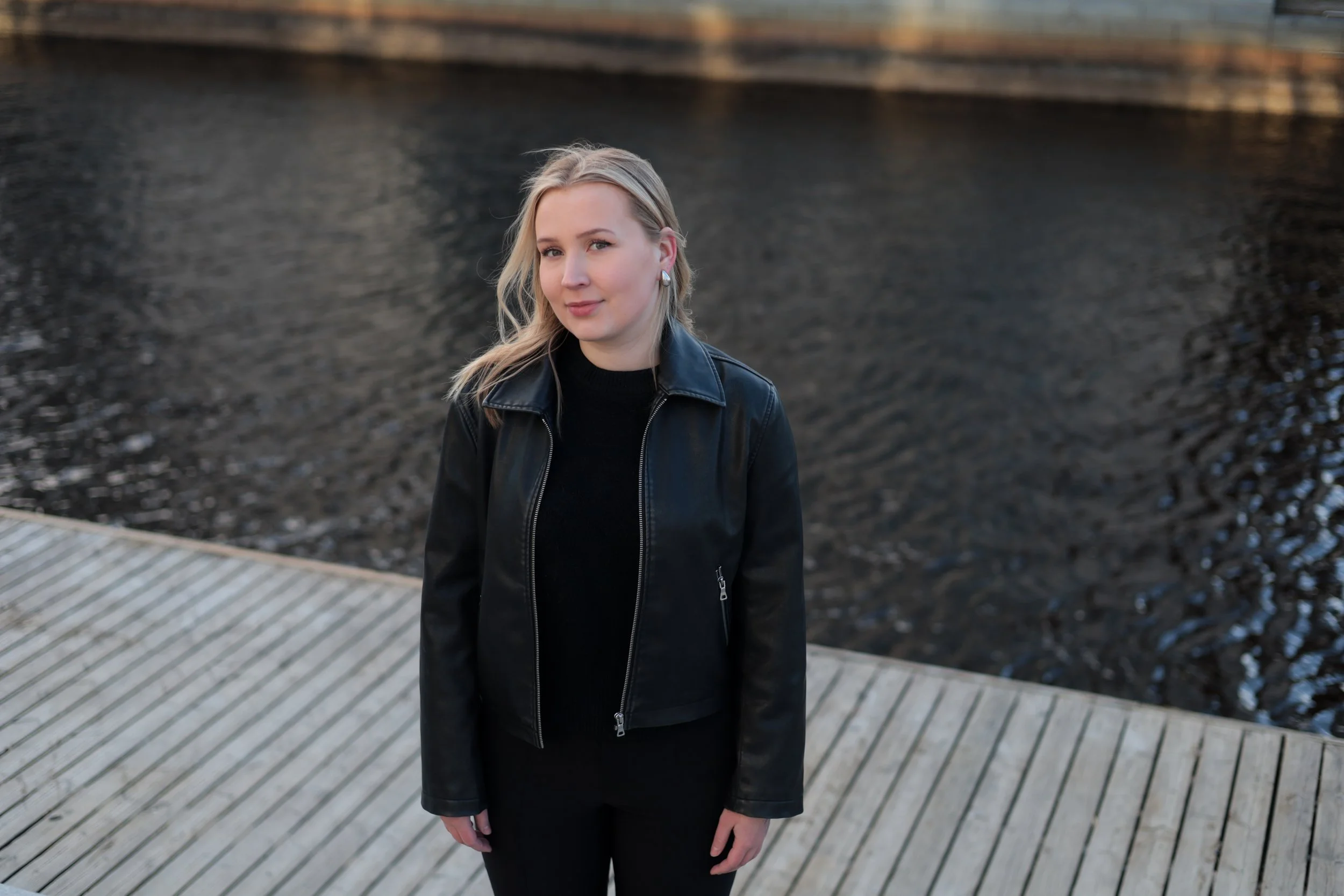 A woman with blonde hair, wearing a black leather jacket, standing on a wooden boardwalk near a body of water.