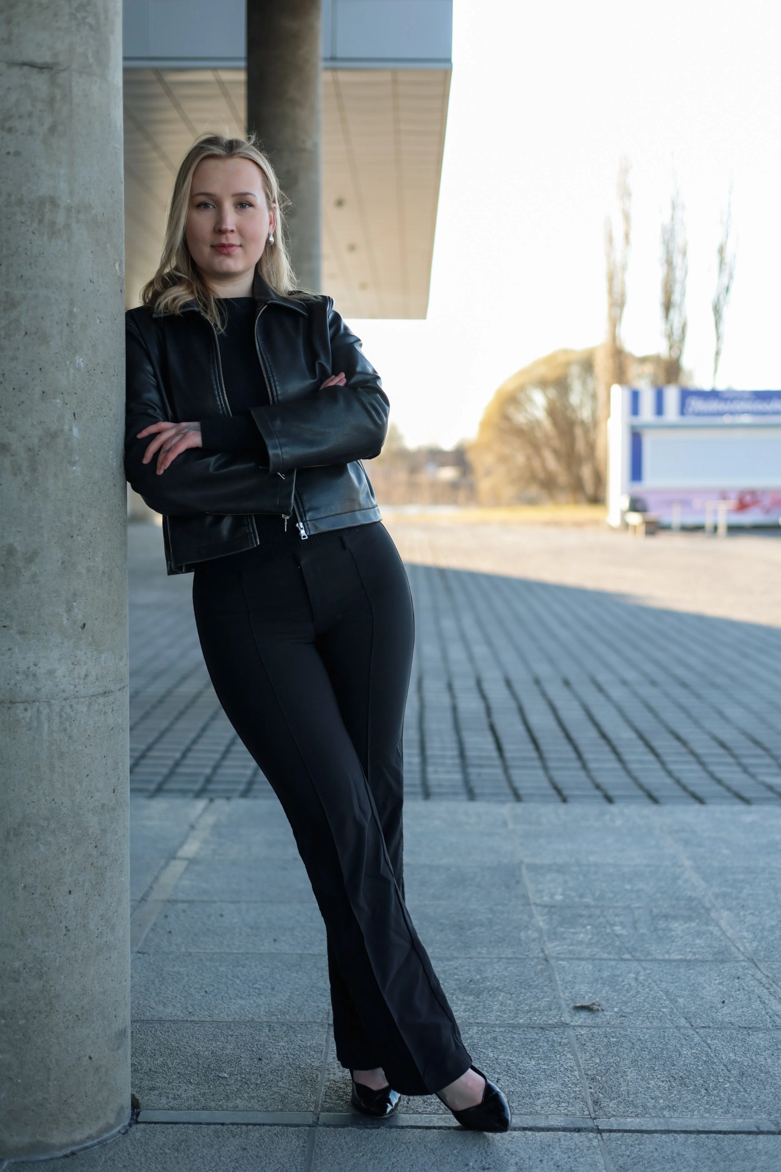 A young woman with blonde hair, wearing a black leather jacket, black pants, and black shoes, is standing outdoors with her arms crossed, leaning against a concrete pillar. She is looking at the camera with a slight smile. In the background, there are blurred trees and a blue and white sign.