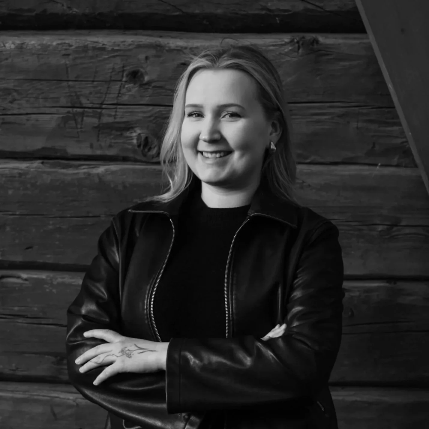 A smiling woman with blonde hair, wearing a black leather jacket and earrings, standing in front of a wooden wall.