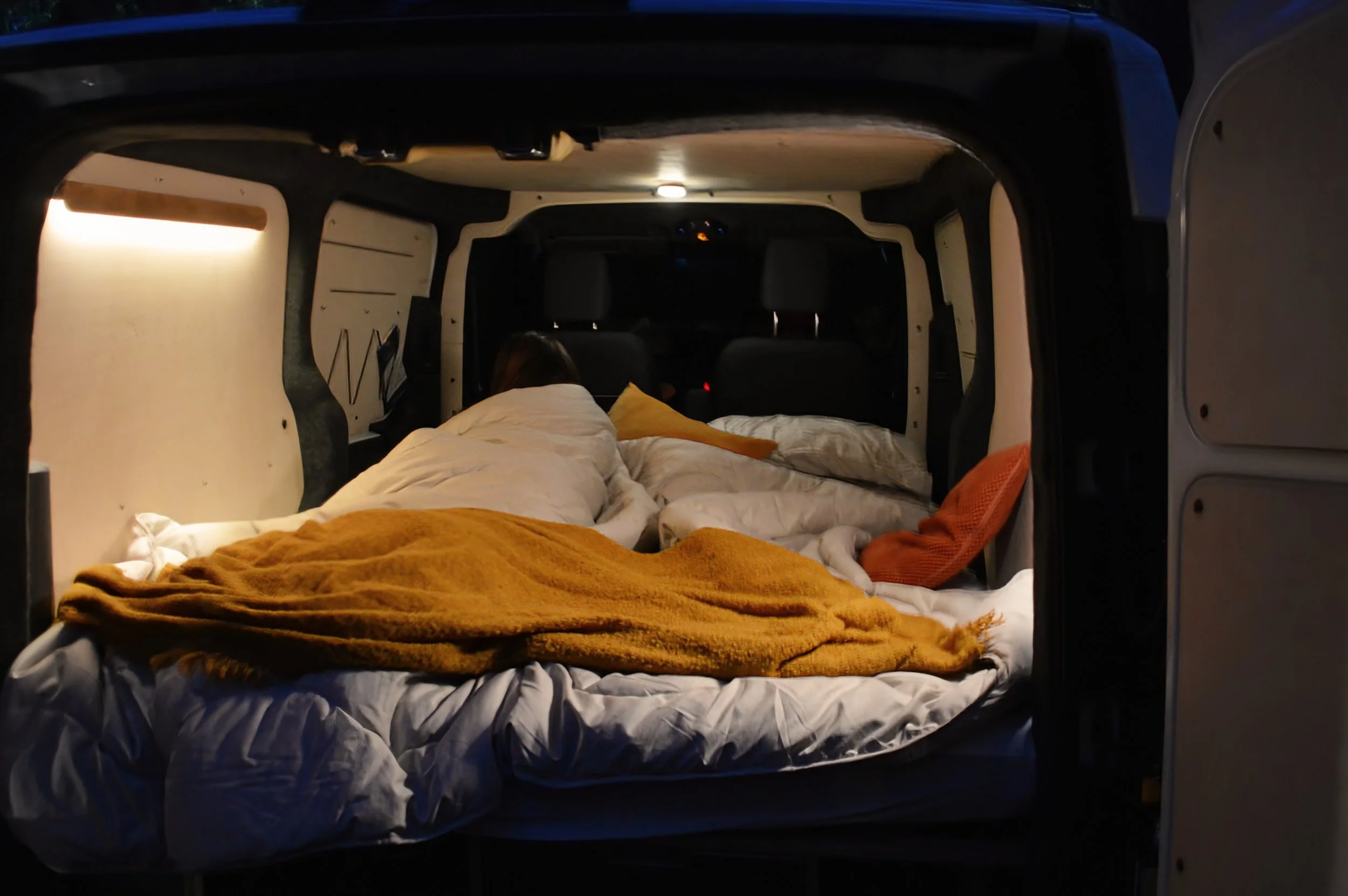 The interior of an electric campervan with warm lighting, equipped for sleeping with blankets and pillows, seen from the rear door open at dusk or night.