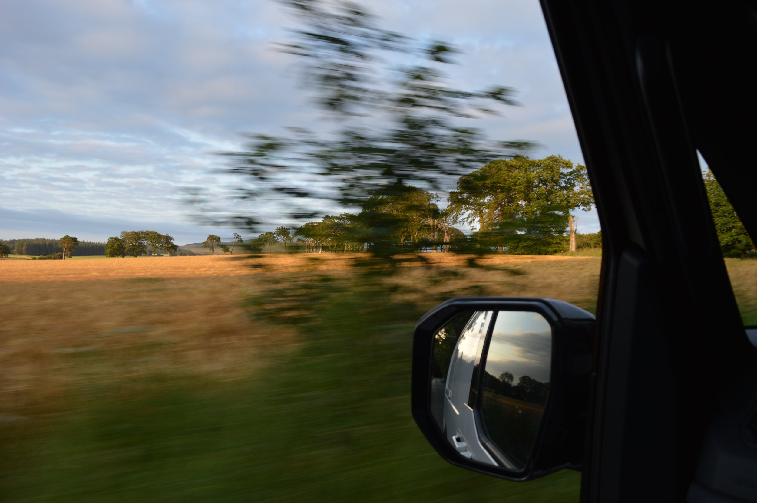 Photo taken from inside a moving vehicle showing a landscape with trees and a blue sky with clouds. The side mirror and part of the vehicle door are visible.
