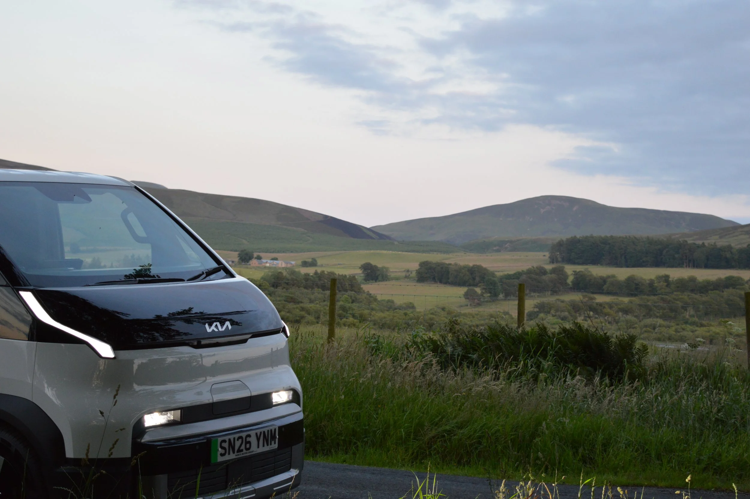 Front view of a white Kia vehicle parked on a grassy roadside with rolling green hills and a cloudy sky in the background.