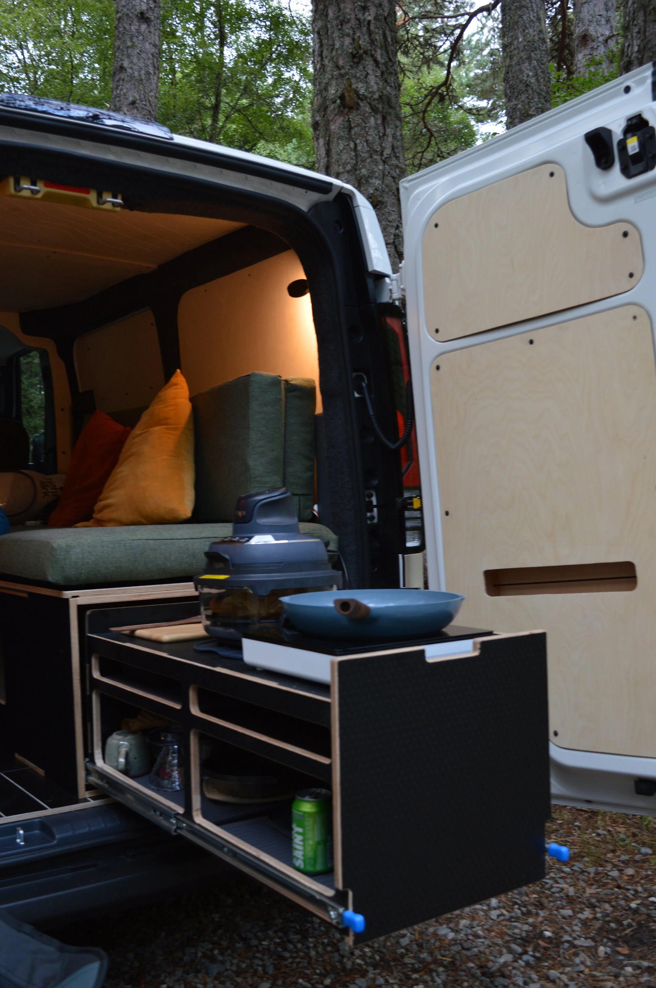 Interior of electric campervan with slide out kitchen, a bed with pillows, and a view of trees outside.