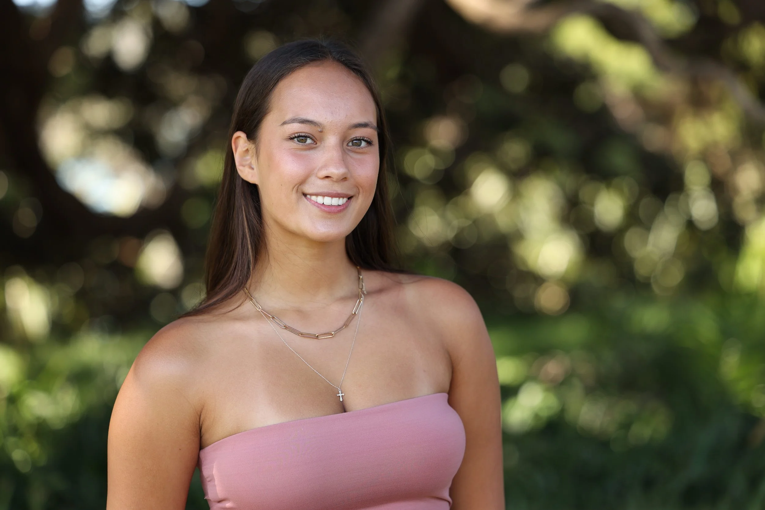 A young woman with long brown hair and a tan complexion smiles outdoors. She is wearing a pink strapless top and layered necklaces, including one with a small cross. The background is blurred with green trees and sunlight.