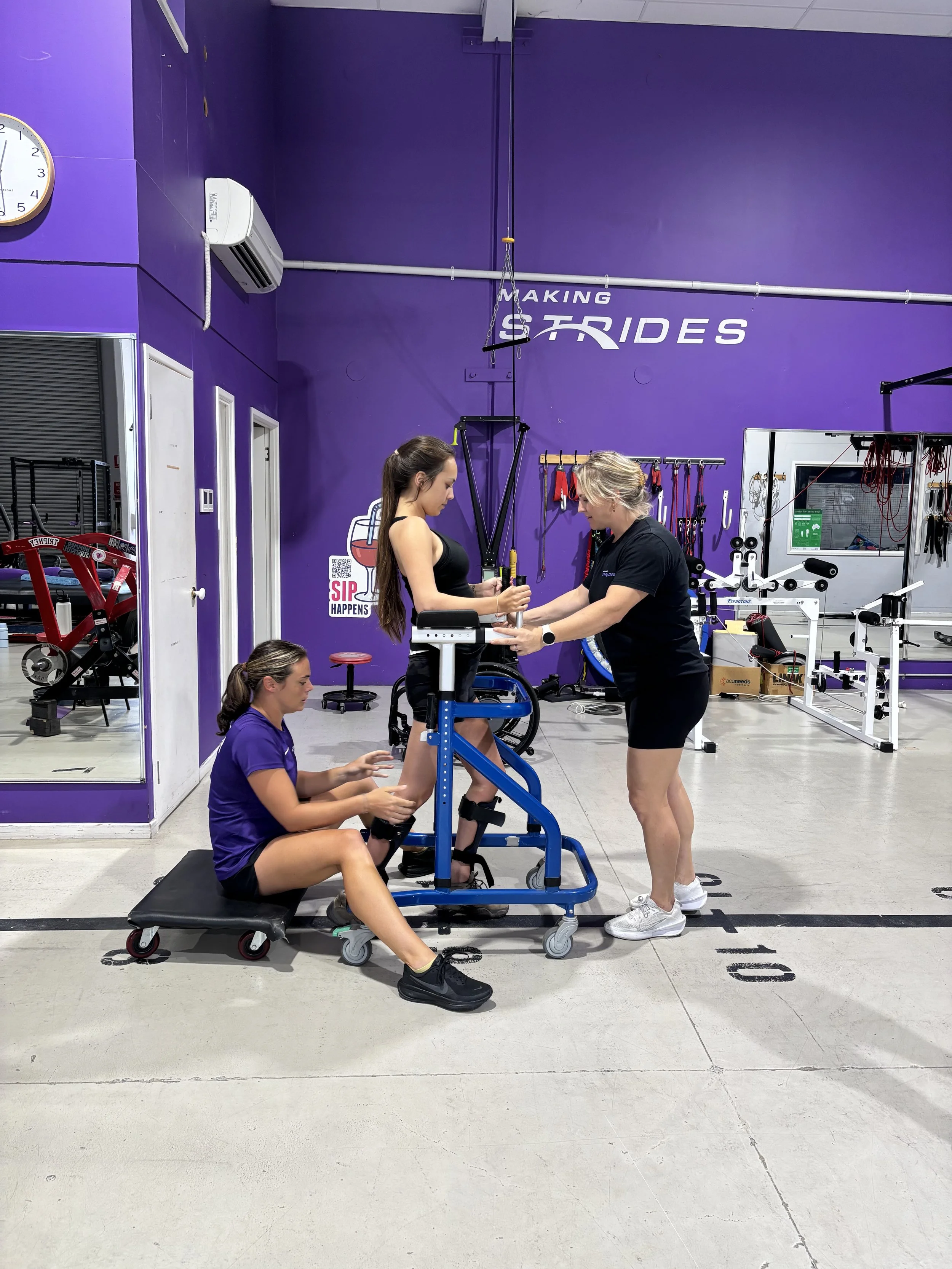 A gym with purple walls where two women are doing wrist curls on a blue wrist curl resistance machine, one woman is standing and the other is sitting on a bench. Another woman is sitting on the floor nearby, looking at her phone. Fitness equipment and a clock are visible in the background.