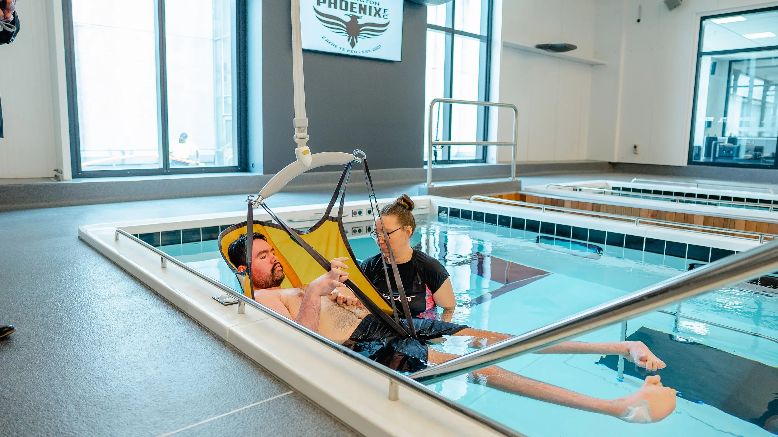 A man is lying in a hospital bed with a bib and a woman is beside him holding his hand, both are in a therapy pool. The pool has a harness attachment with a person's head visible in the harness, assisting in water therapy. Large windows and a sign with a phoenix logo are in the background.