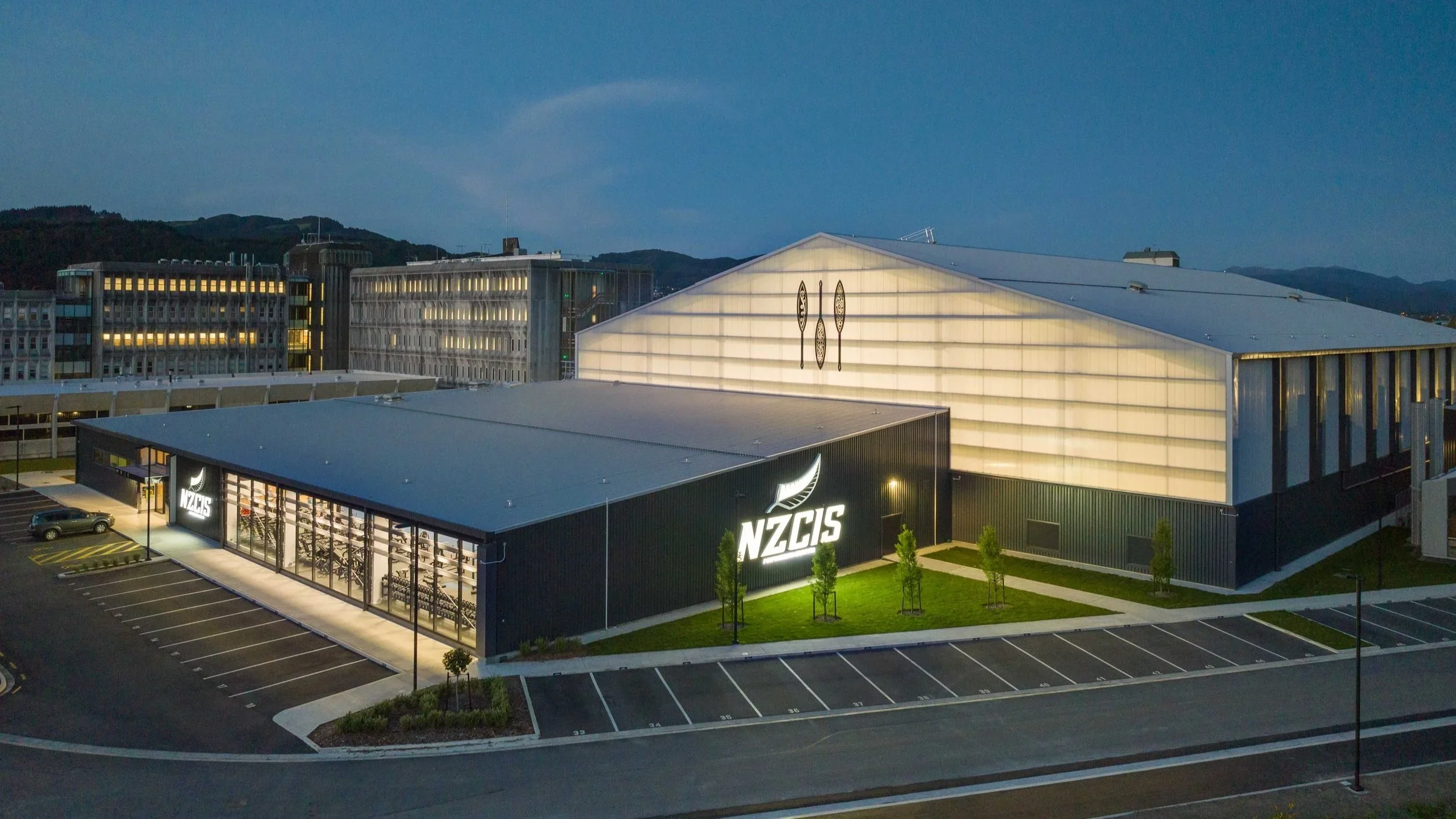A modern building with the NZCIS logo and sports equipment inside, located in a parking lot with trees, against a backdrop of hills and a blue evening sky.