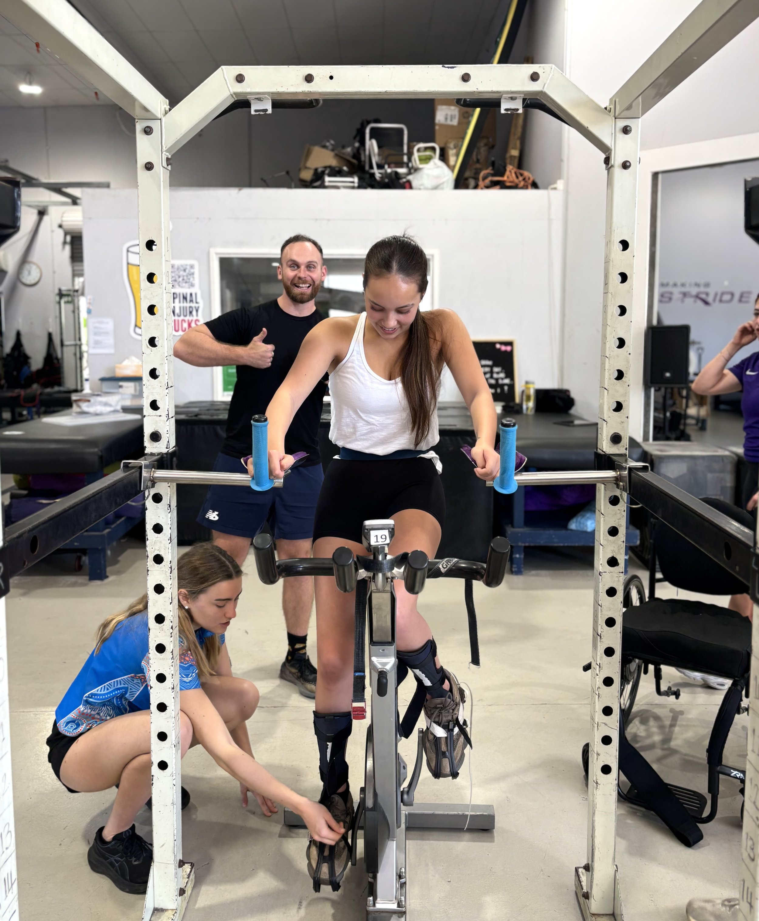 A woman in athletic gear is preparing to ride a stationary exercise bike, with a man giving a thumbs-up in the background. Two women are crouched on the floor, adjusting her shoe or gear, in a gym or fitness center setting.