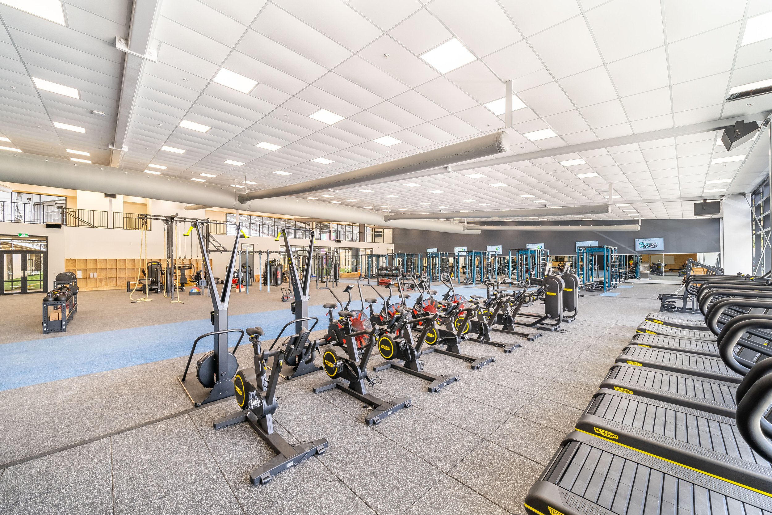 An empty gym with various exercise machines and equipment, including stationary bikes and weights, in a spacious room with high ceilings and large windows.