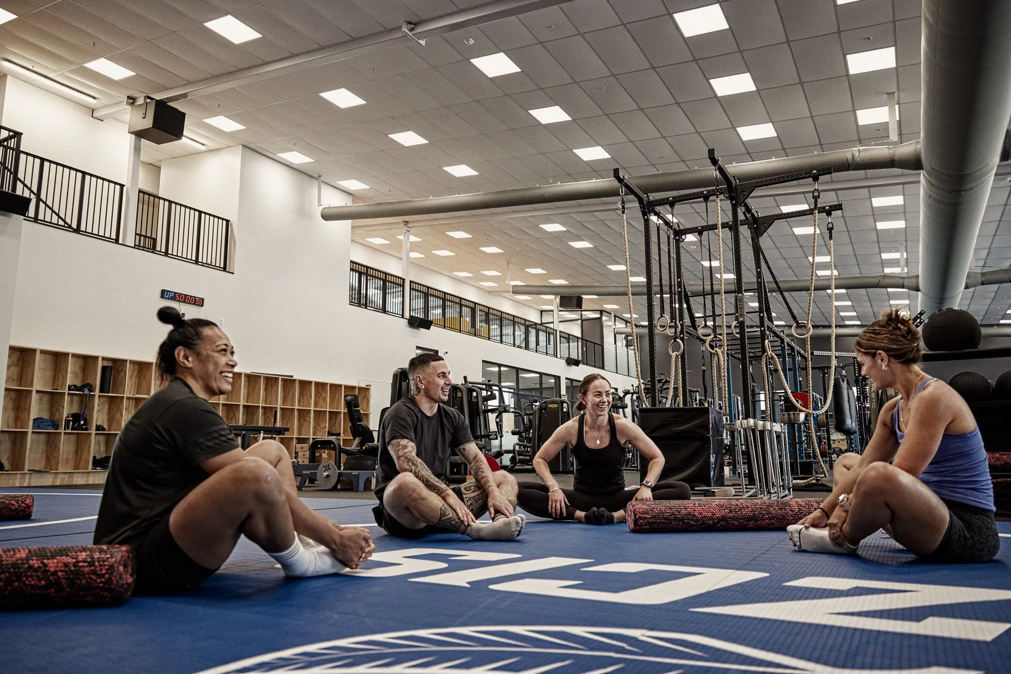 A group of five people sitting on yoga mats in a gym, smiling and engaging in conversation. The gym has a high ceiling, large windows, and fitness equipment such as rings, ropes, and weight machines in the background.